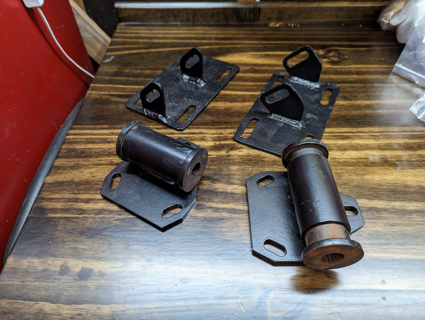 Four black metal brackets from The Ugly Truckling KIT LS Swap Engine Mounts for 1998-2005 Chevy Blazer, GMC Jimmy, and two cylindrical bushings with rusted ends are displayed on a wooden surface. The brackets feature angled tabs with holes.