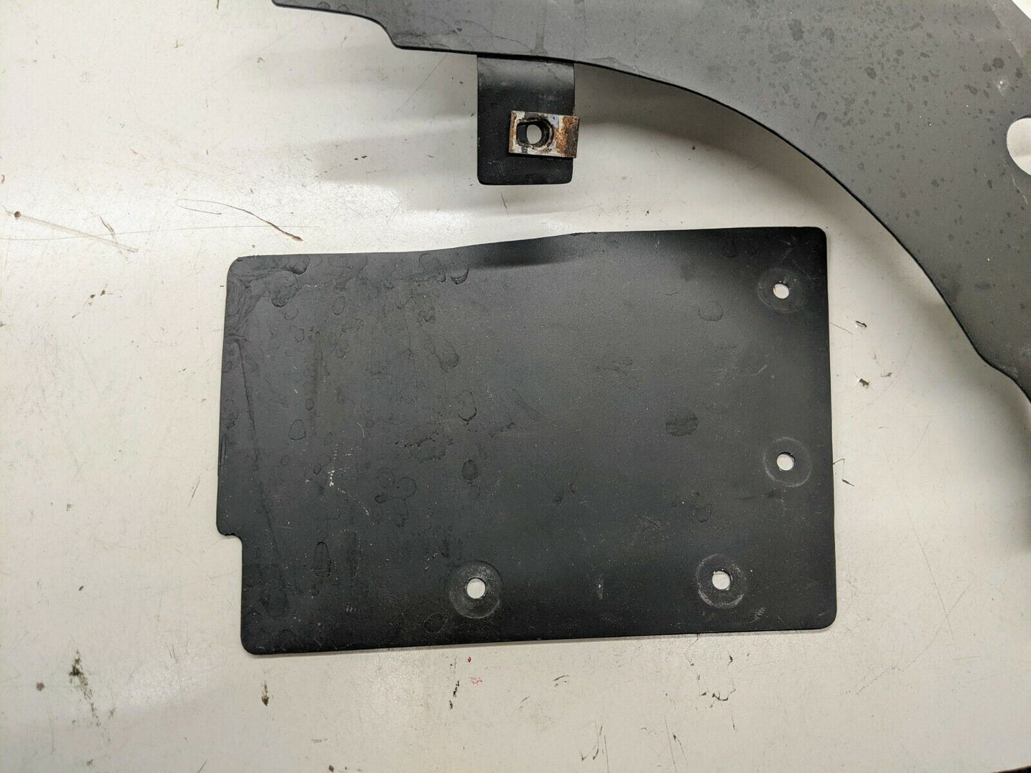 A black metal plate with four holes and a curved gray custom bracket with a rusted bolt, both by The Ugly Truckling, are shown—parts for the 1995–2005 Chevy S10 Blazer LS Swap Cold Air Box Custom Fabricated Factory Look setup.