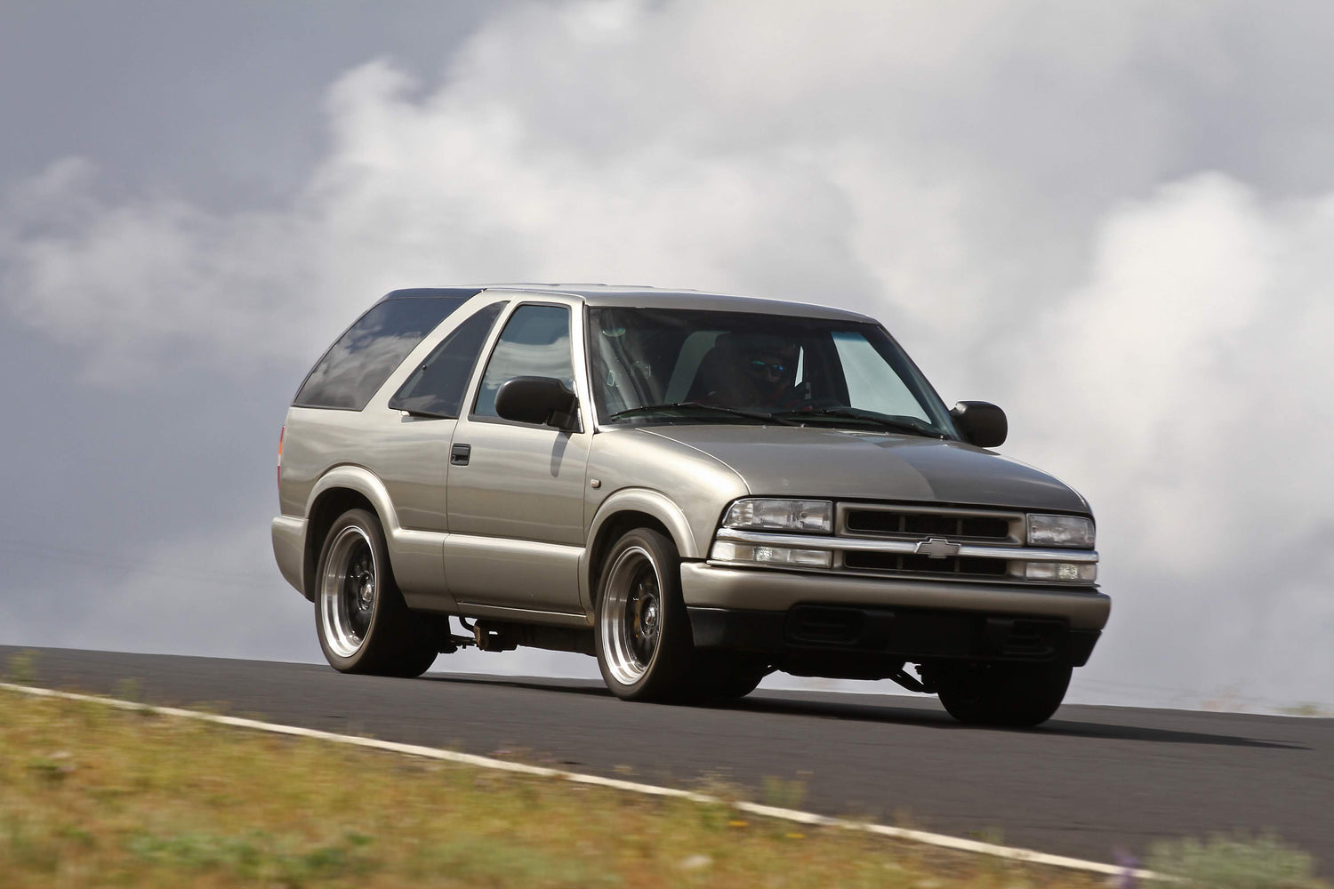 A silver two-door SUV, likely a Chevrolet, drives on a paved road with grass along the edge and a cloudy sky in the background.