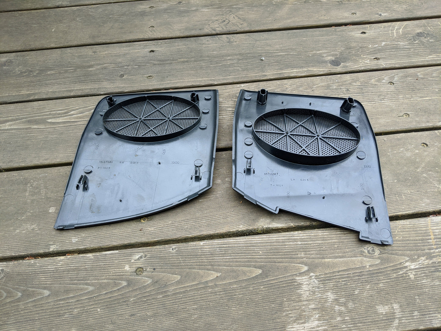 An OEM pair of General Motors dark gray dash speaker grille covers (LH & RH) for 1995-1997 Chevrolet S10, Blazer, and more are shown side by side on a wooden deck, featuring rectangular shape, rounded edges, mounting holes, and pegs.
