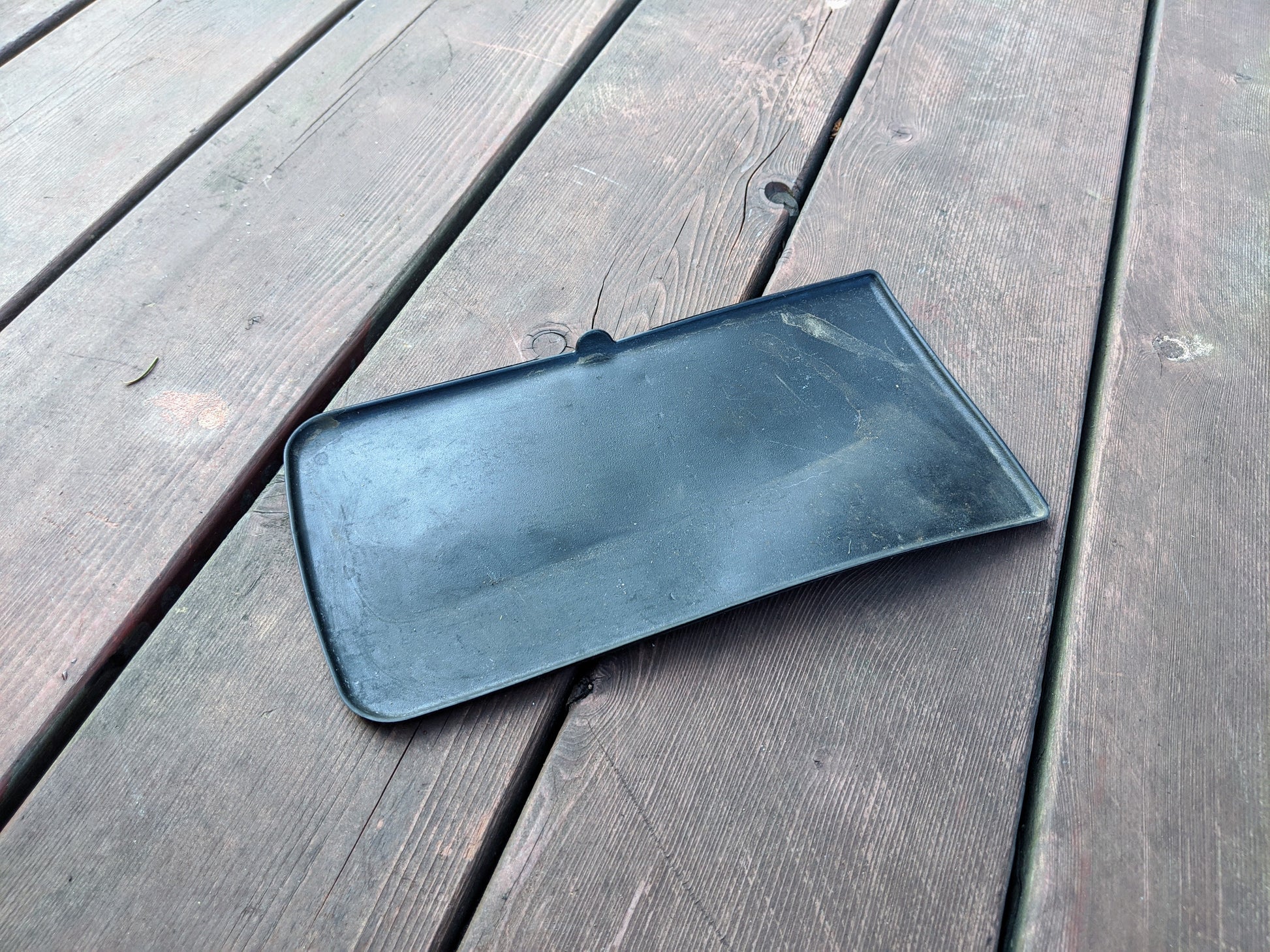 A black OEM Center Console Storage Bin Rubber Mat Liner by General Motors for 2003-2006 Chevy Silverado/GMC Sierra rests on a weathered wooden deck, showing signs of use with smudges and dirt on its surface.