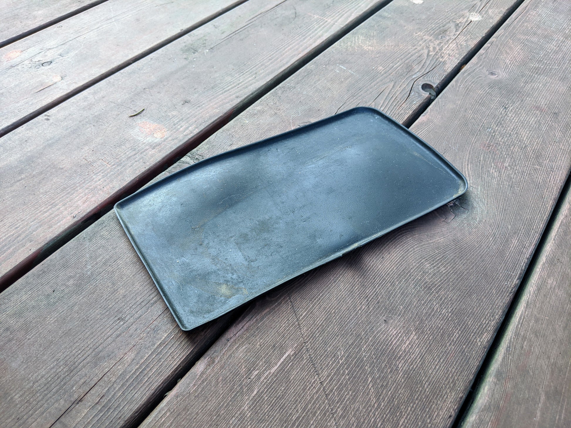 A black, rectangular OEM Center Console Storage Bin Rubber Mat Liner by General Motors for 2003-2006 Chevy Silverado/GMC Sierra rests on a weathered deck, showing some warping, scuffs, and discoloration from use.