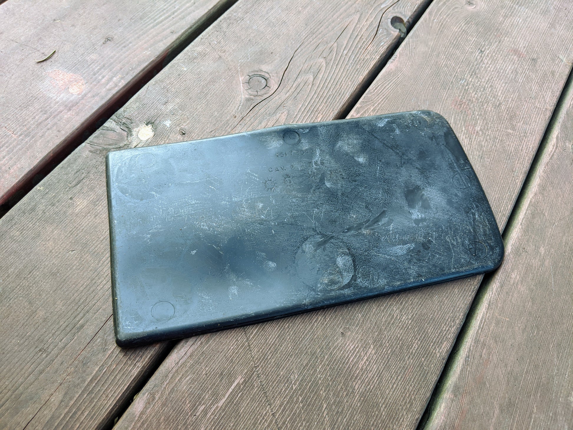 A weathered, rectangular black plastic object with rounded corners lies on wooden planks. Slightly scratched and scuffed, it resembles an OEM Center Console Storage Bin Rubber Mat Liner by General Motors for 2003-2006 Chevy Silverado GMC Sierra.