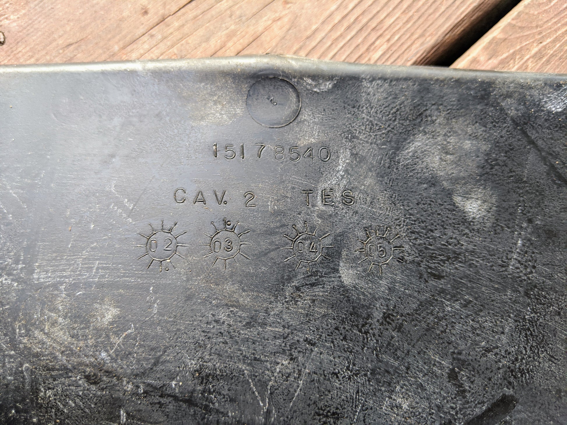 Close-up of a black General Motors OEM Center Console Storage Bin Rubber Mat Liner, embossed with text and starburst symbols, designed for 2003-2006 Chevy Silverado, GMC Sierra, and more—shown on a wooden background.