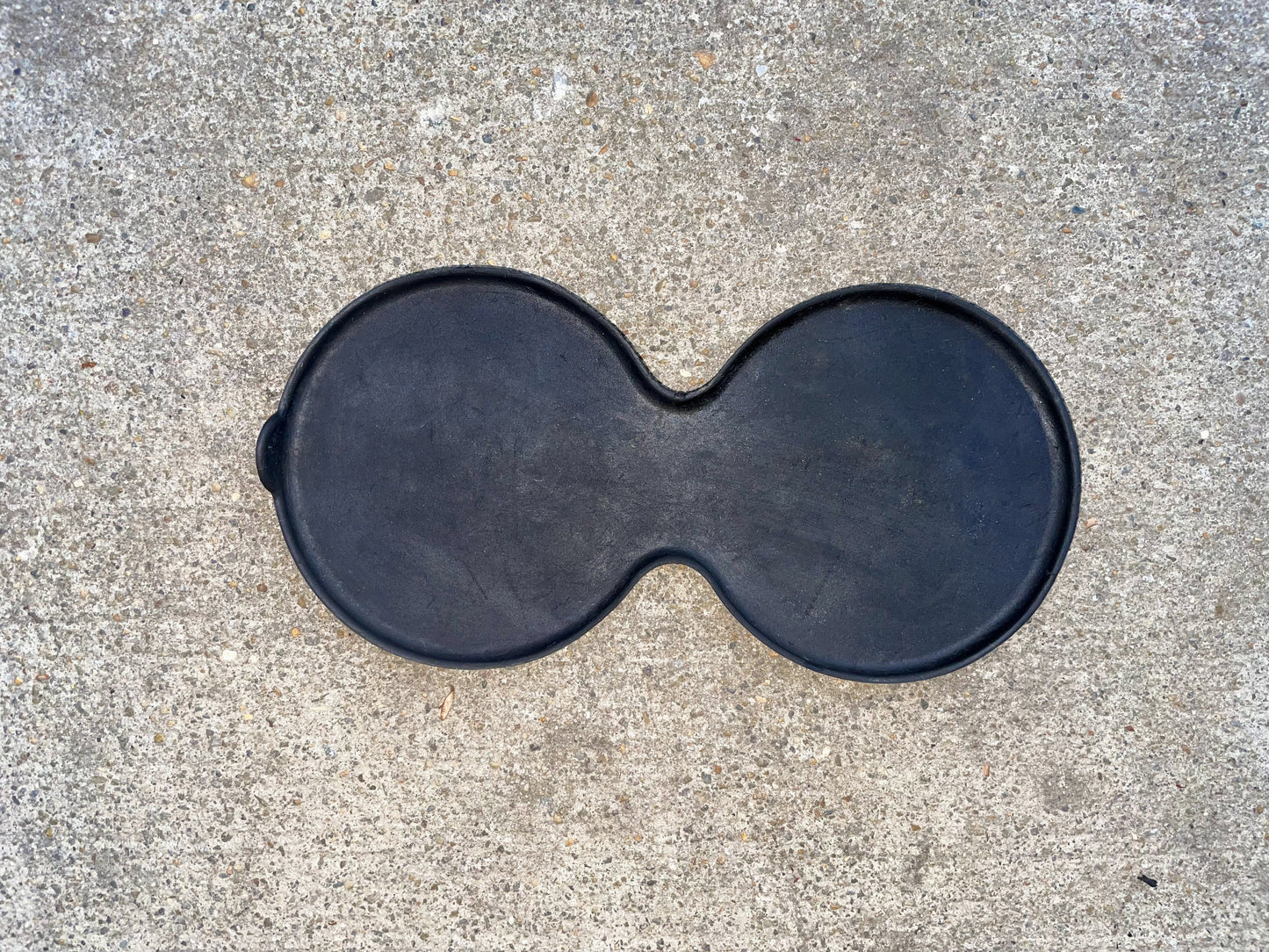 A black, figure-eight-shaped griddle rests on rough light gray concrete, resembling the General Motors OEM Center Console Cup Holder Bin Rubber Mat Liner for 2003-2006 Chevy Silverado and GMC Sierra.
