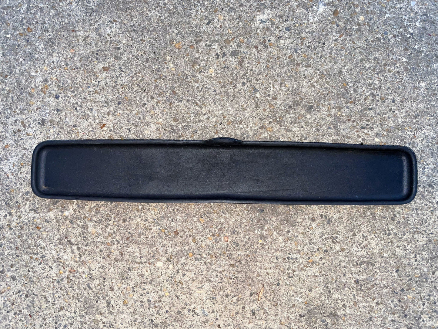 A black, rectangular rubber mat with rounded corners lies on speckled concrete; it resembles the General Motors OEM Center Console Cup Holder Central Storage Bin Rubber Mat Liner for 2003-2006 Chevy Silverado, GMC Sierra, and similar models.