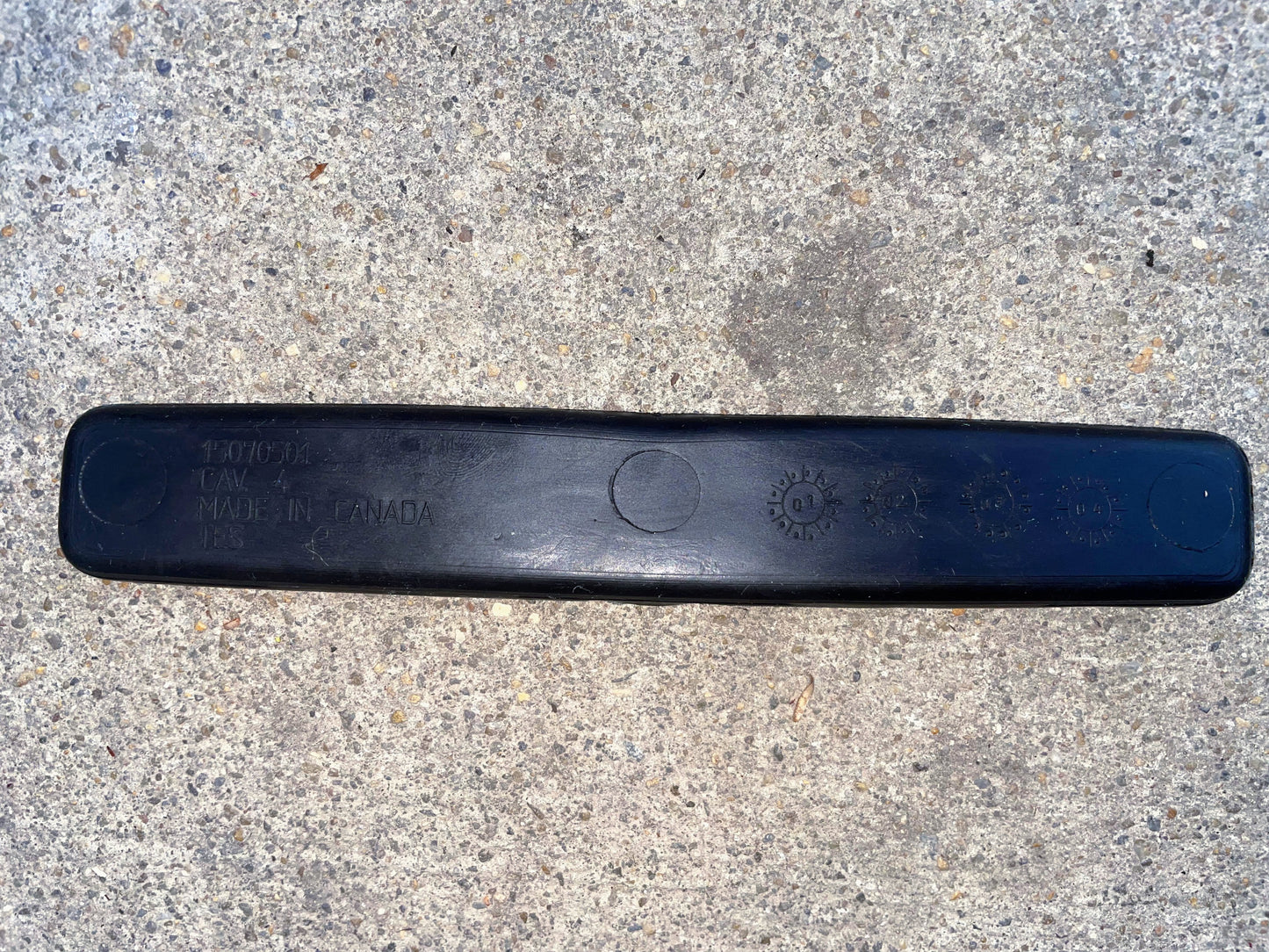 A black General Motors OEM Center Console Cup Holder Storage Bin Rubber Mat Liner for 2003-2006 Chevy Silverado/GMC Sierra features embossed text, including MADE IN CANADA and symbols, resting on rough light gray concrete.