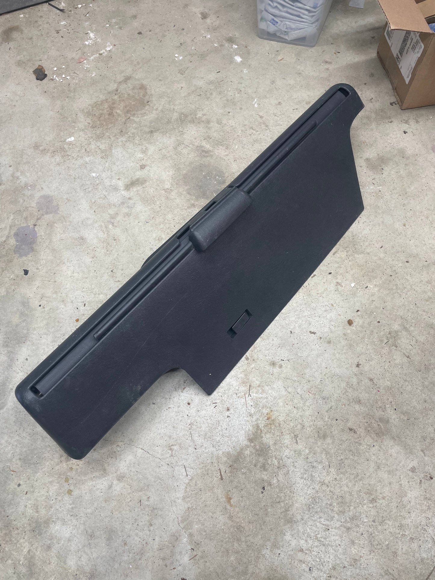 A General Motors OEM Retractable Rear Cargo Shade Jack Cover in Graphite for 1995-2005 Chevy Blazer GMC Jimmy stands upright on a concrete floor, with storage boxes and other objects in the background.