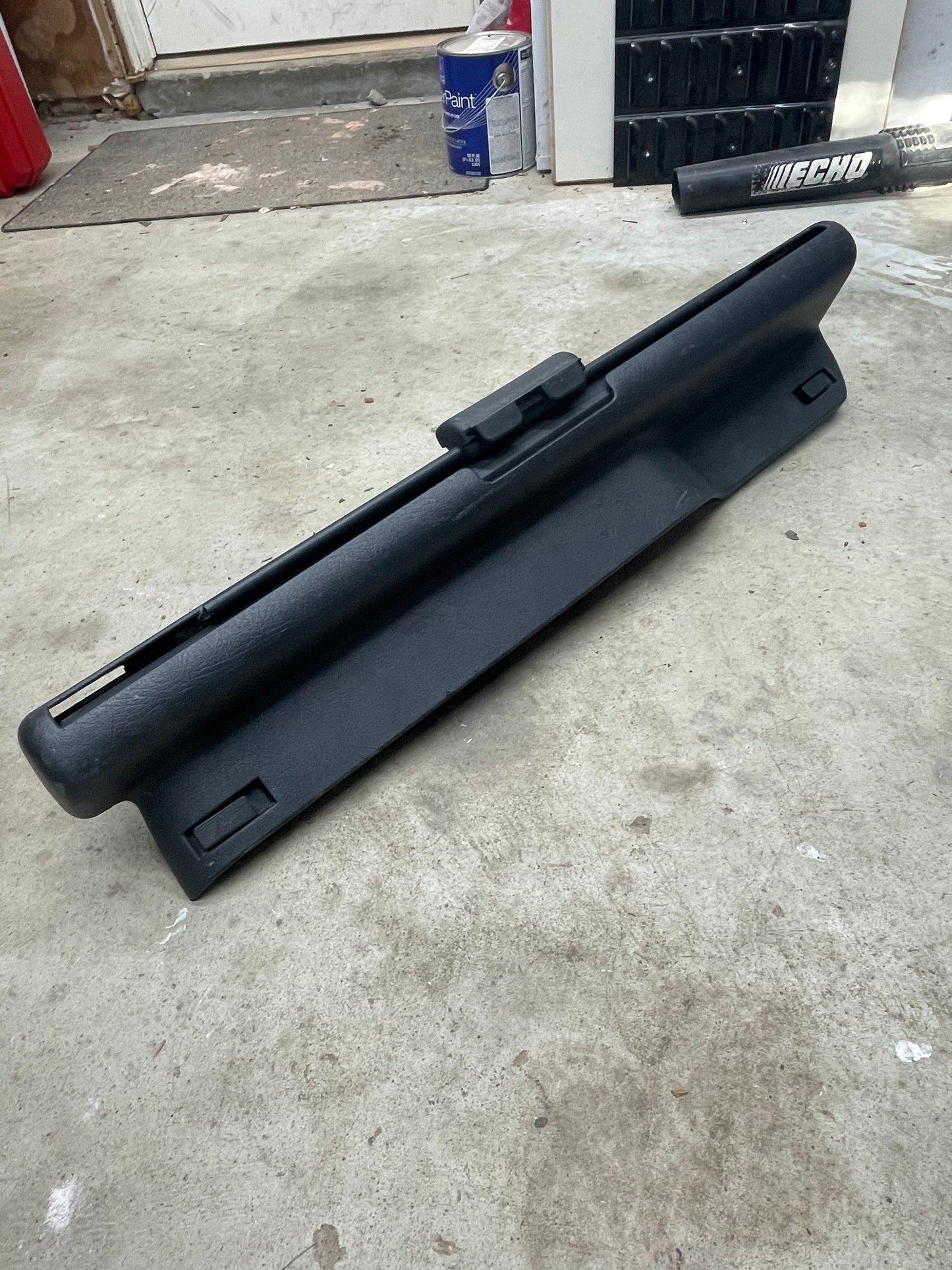 A General Motors OEM Retractable Rear Cargo Shade Jack Cover in Graphite for 1995-2005 Chevy Blazer/GMC Jimmy sits on a concrete garage floor, with tools, a paint can, and other items visible in the background.