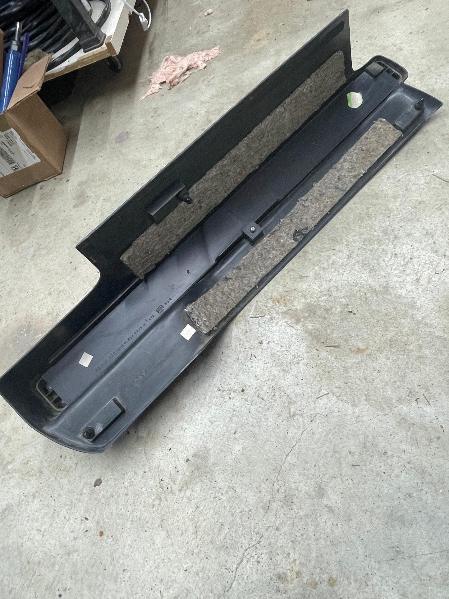 A General Motors OEM Retractable Rear Cargo Shade Jack Cover in graphite for 1995-2005 Chevy Blazer and GMC Jimmy lies on a concrete floor, surrounded by cardboard boxes and various workshop supplies.