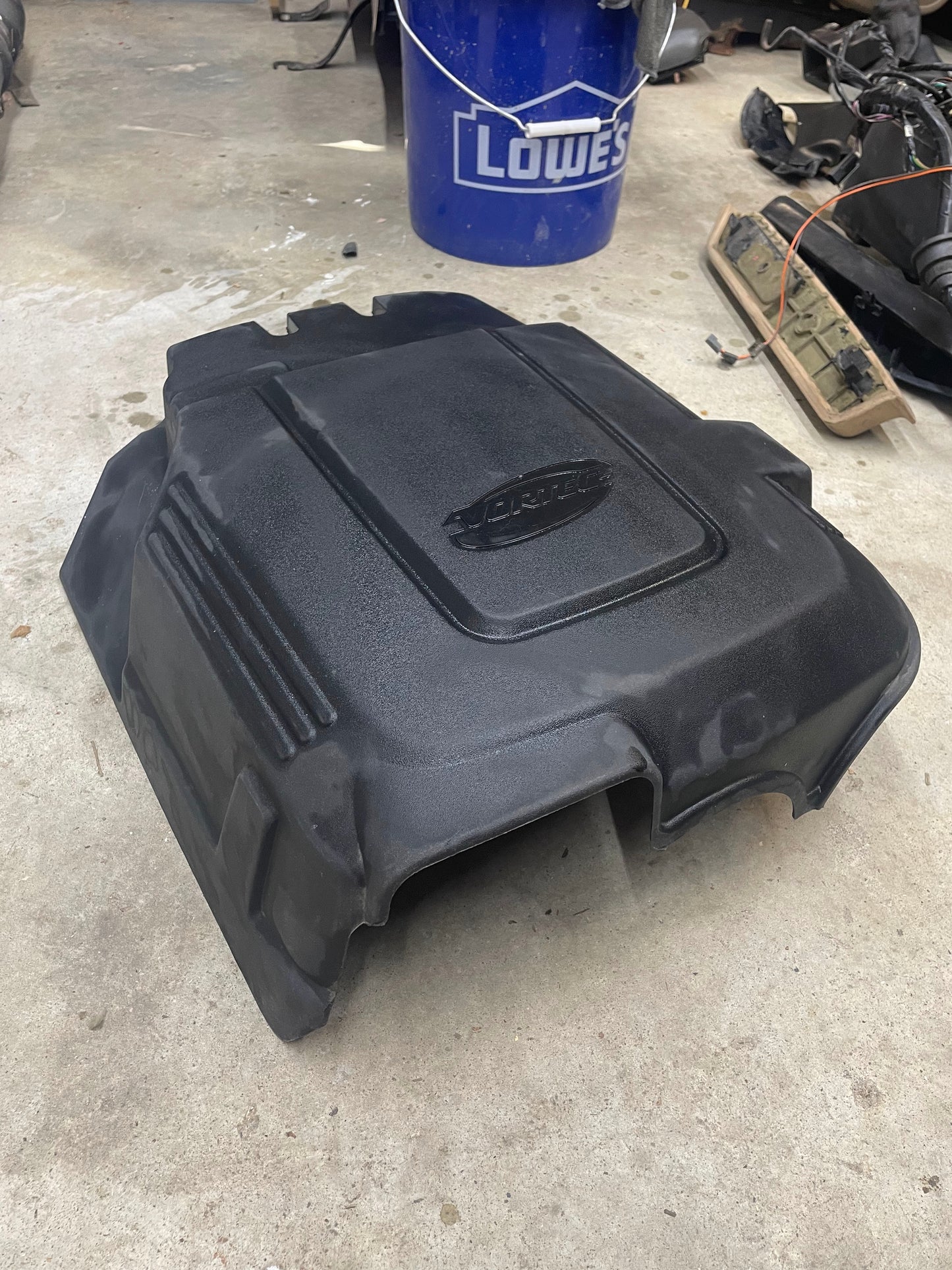 A General Motors OEM Vortec Engine Intake Cover for 2007-2019 GM Trucks and LS swaps, made of black plastic, rests on a concrete floor. A blue Lowes bucket and assorted garage tools are visible in the background.