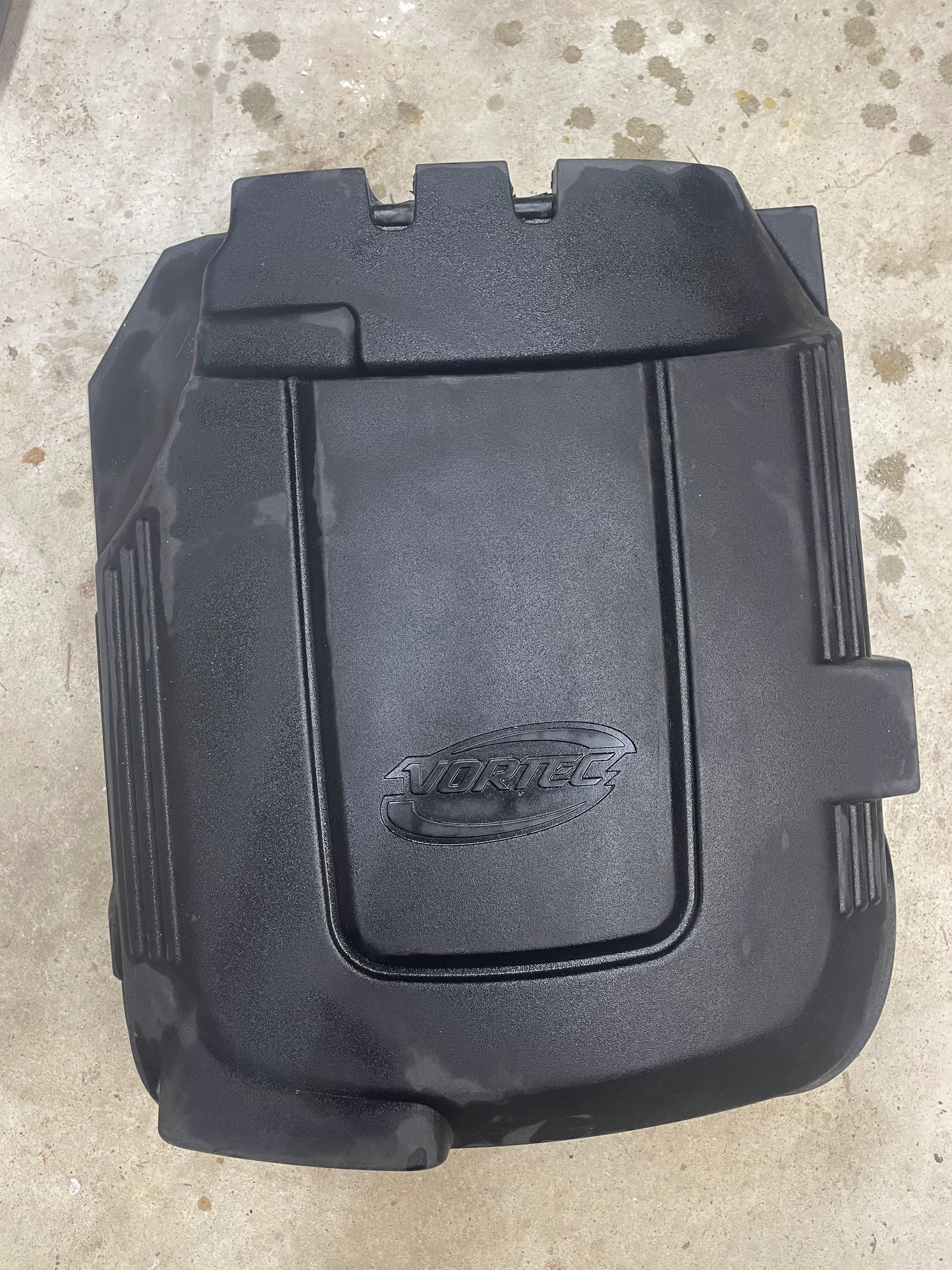 A General Motors OEM Vortec Engine Intake Cover for 2007-2019 GM Trucks and LS Swaps, made of black plastic and embossed with VORTEC, is shown resting on a stained concrete surface.