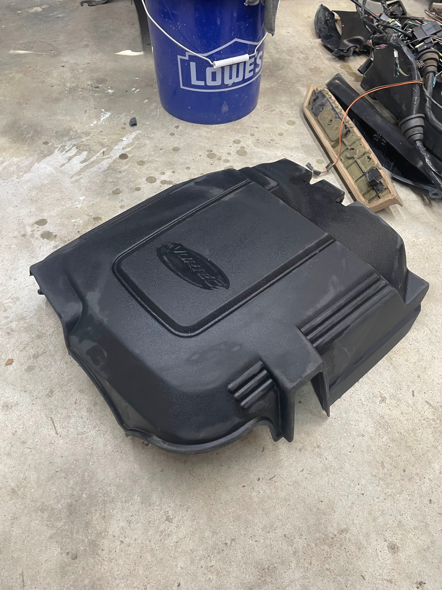 An OEM Vortec Engine Intake Cover by General Motors for 2007-2019 GM Trucks and LS swaps rests on a concrete floor, with a blue Lowe’s bucket and various car parts and tools nearby.