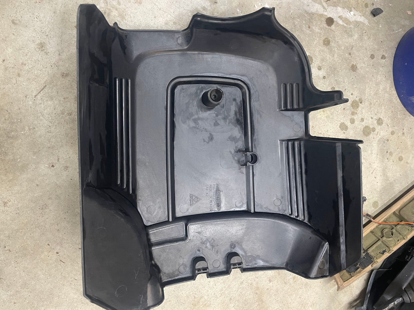 A large black plastic General Motors OEM Vortec Engine Intake Cover for 2007-2019 GM Trucks and LS swaps, with grooves, raised sections, curved edges, and a central round protrusion, is placed on a concrete floor.