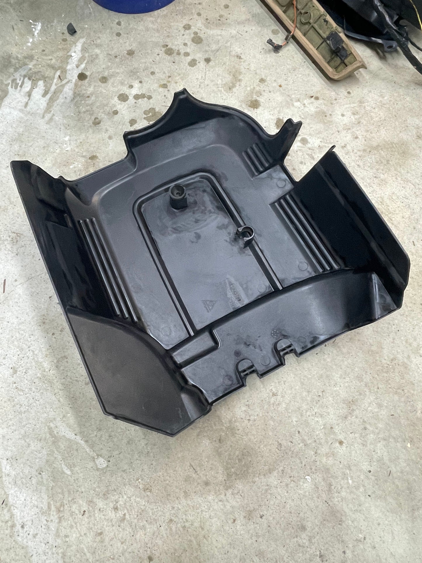 A General Motors OEM Vortec Engine Intake Cover for 2007-2019 GM Trucks and LS Swaps, made of black plastic with raised edges and ridges, rests on a concrete floor with visible dirt and water stains nearby.