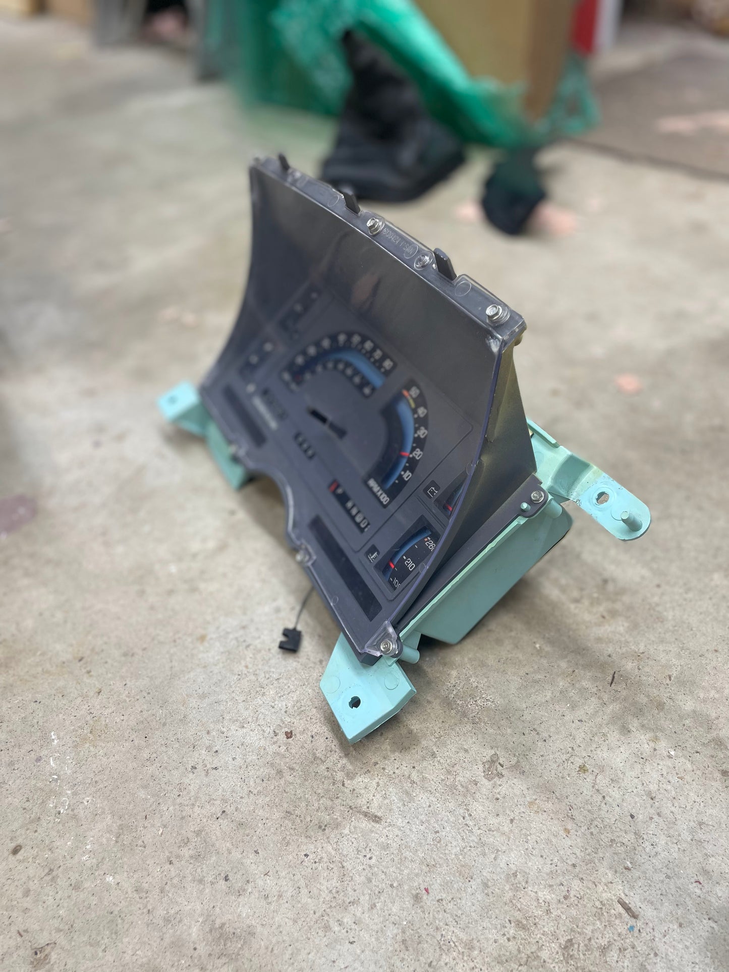 A LOW Mile 72K OEM General Motors instrument cluster with mechanical speedo, tachometer, and PRNDL for 1986-1989 Chevy S10 Blazer rests on concrete, featuring a clear cover, green mounts, and small loose parts.