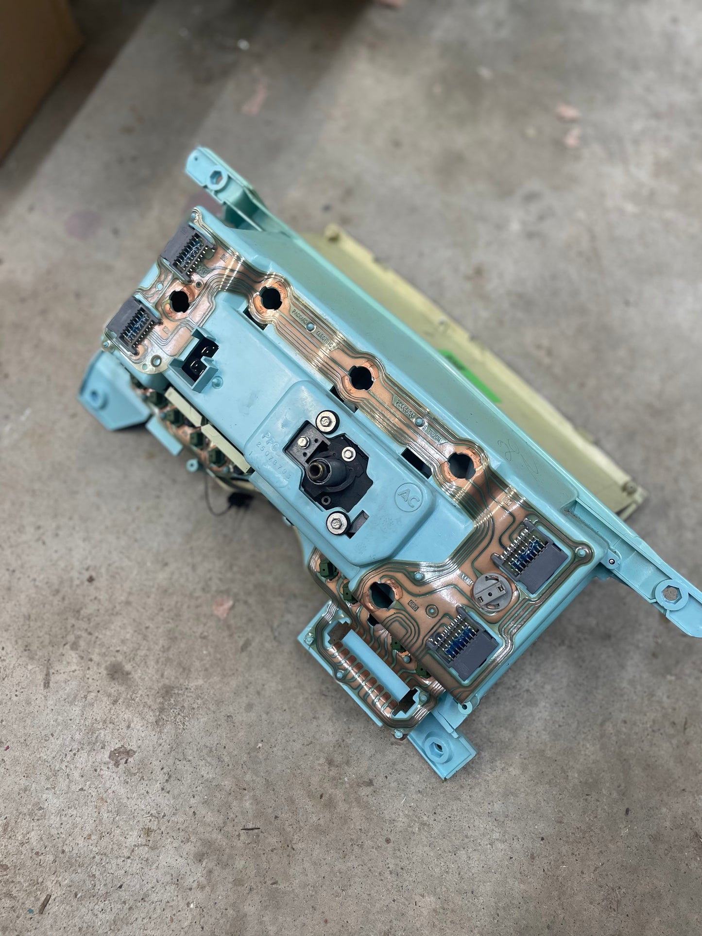 A General Motors LOW Mile 72K OEM Instrument Cluster with mechanical speedo, tachometer, and PRNDL for 1986-1989 Chevy S10 Blazer rests on a concrete floor, its teal frame showing signs of wear and discoloration.