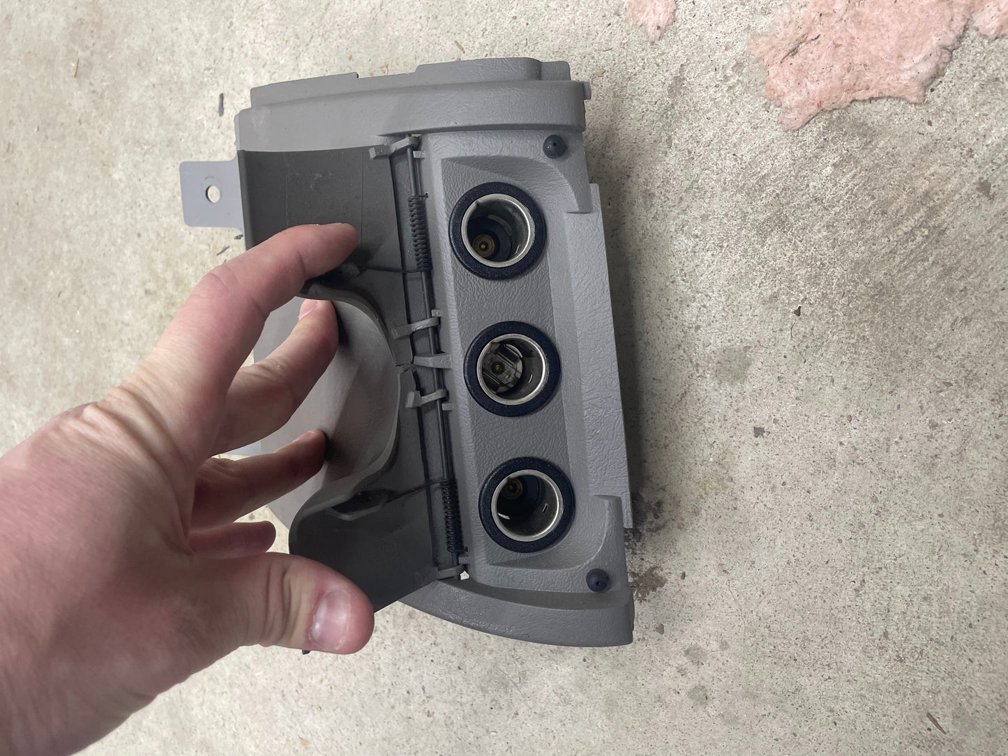 A hand holds a General Motors OEM 3 Port 12V Aux Panel Ash Tray Delete in light gray, designed for 1998-2000 GMC Envoy, featuring three circular openings and placed on a cement floor.