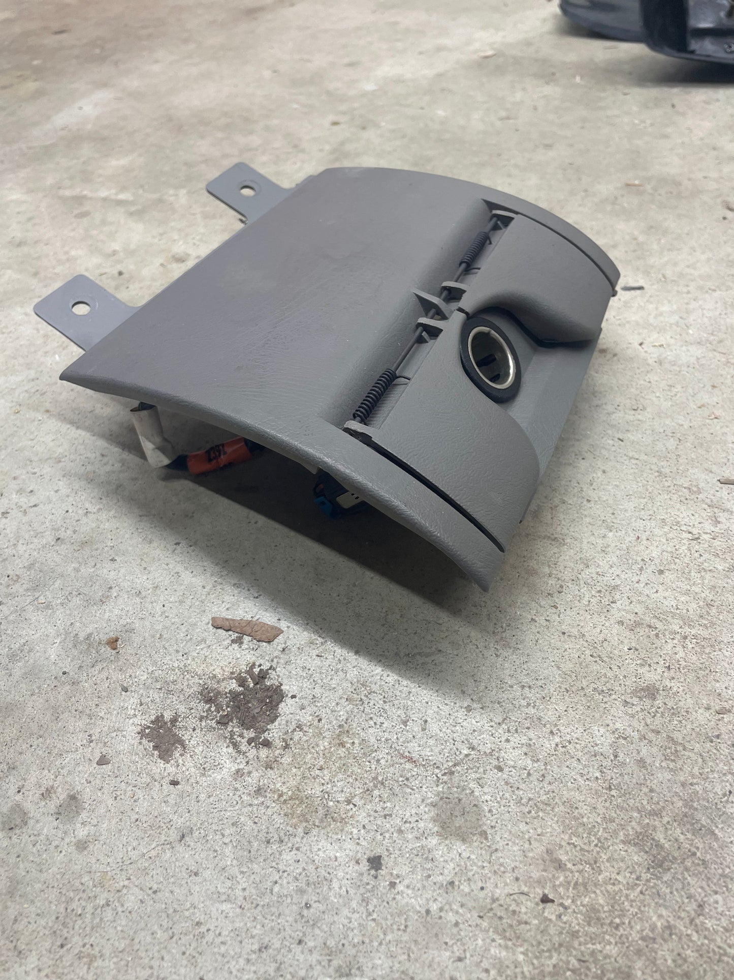 A light gray plastic OEM 3 Port 12V Aux Panel Ash Tray Delete from a 1998-2000 GMC Envoy by General Motors lies detached on a concrete floor, showing hinges, front keyhole, and partially exposed wiring underneath.