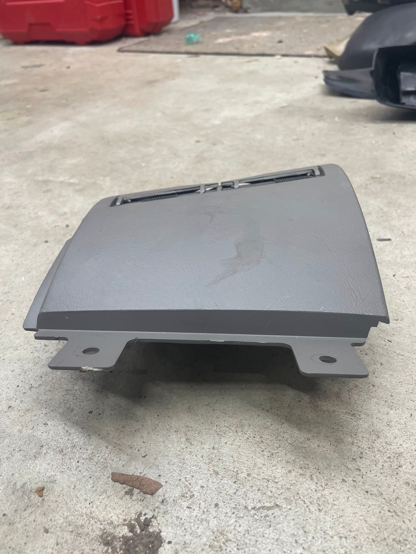 A General Motors OEM 3 Port 12V Aux Panel Ash Tray Delete in Light Gray, from a 1998-2000 GMC Envoy, rests on a concrete floor with tools and items blurred in the background.