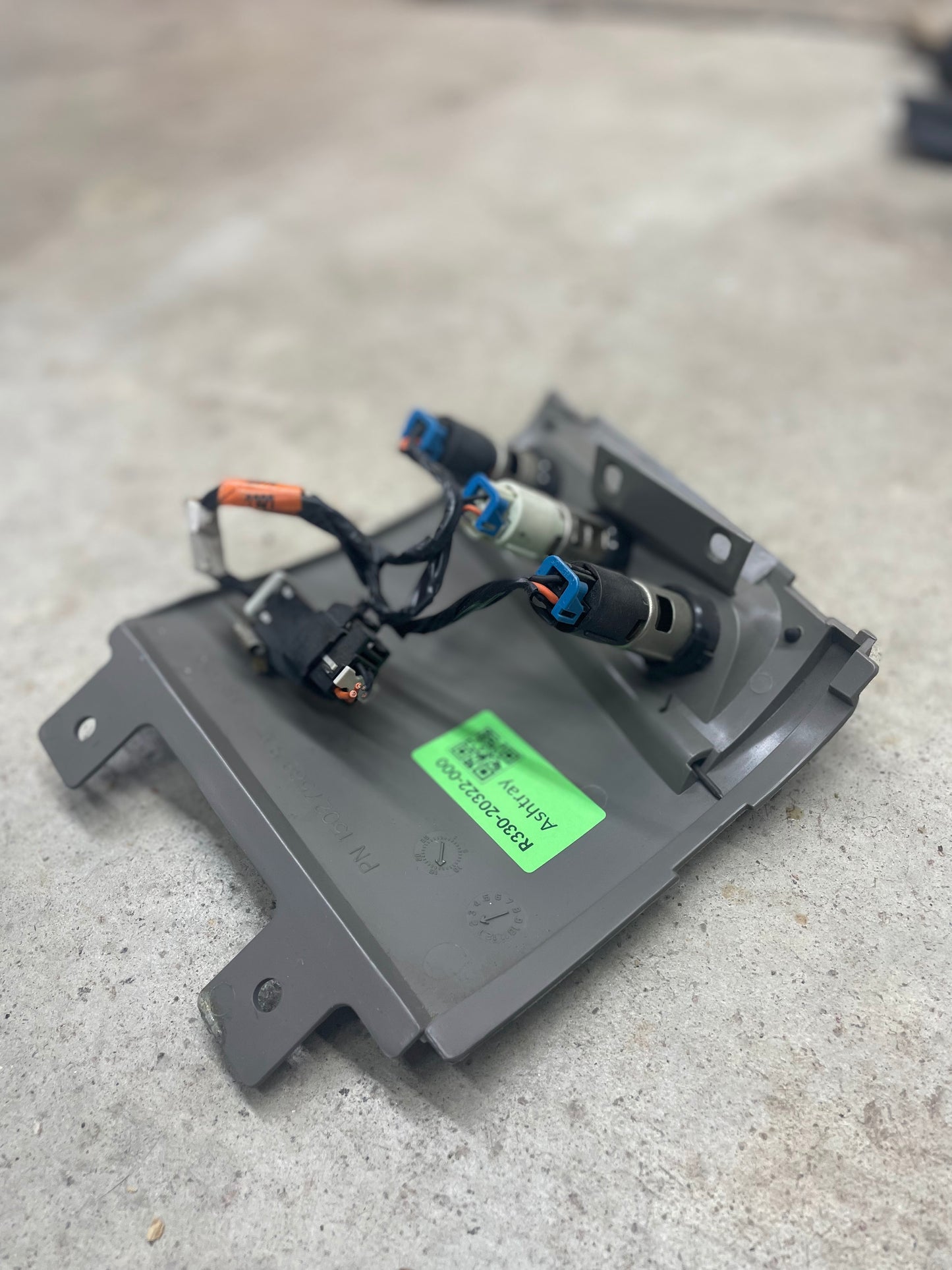 A General Motors OEM 3 Port 12V Aux Panel Ash Tray Delete in Light Gray, from a 1998-2000 GMC Envoy, with three electrical connectors and wires, rests on concrete. A green label with black text is visible on the part.
