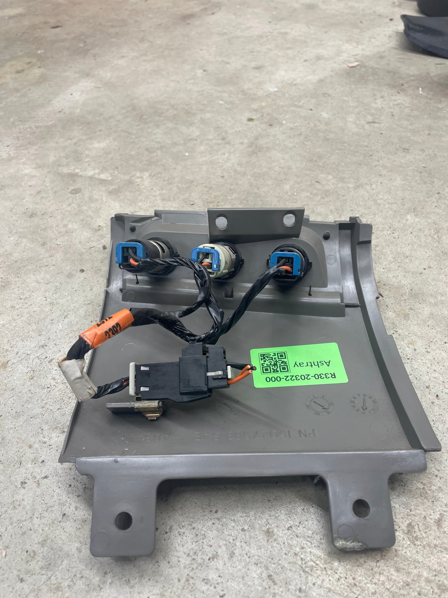 A General Motors OEM 3 Port 12V Aux Panel Ash Tray Delete in Light Gray from a 1998-2000 GMC Envoy, featuring three connectors, black wires, an orange-tagged cable, and a green barcode label, shown on concrete.