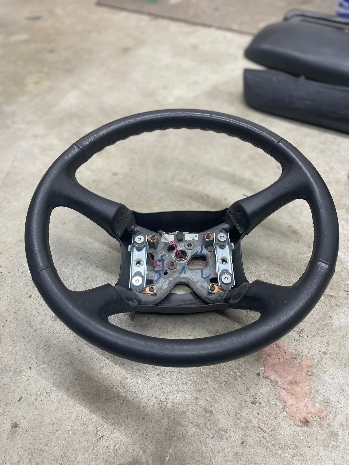 An OEM Leather Steering Wheel by General Motors for 1999-2005 GM Chevy Silverado, S10, and more is shown without its central cover, revealing the mounting hardware and wiring, lying on a concrete floor with a dark cushion behind.