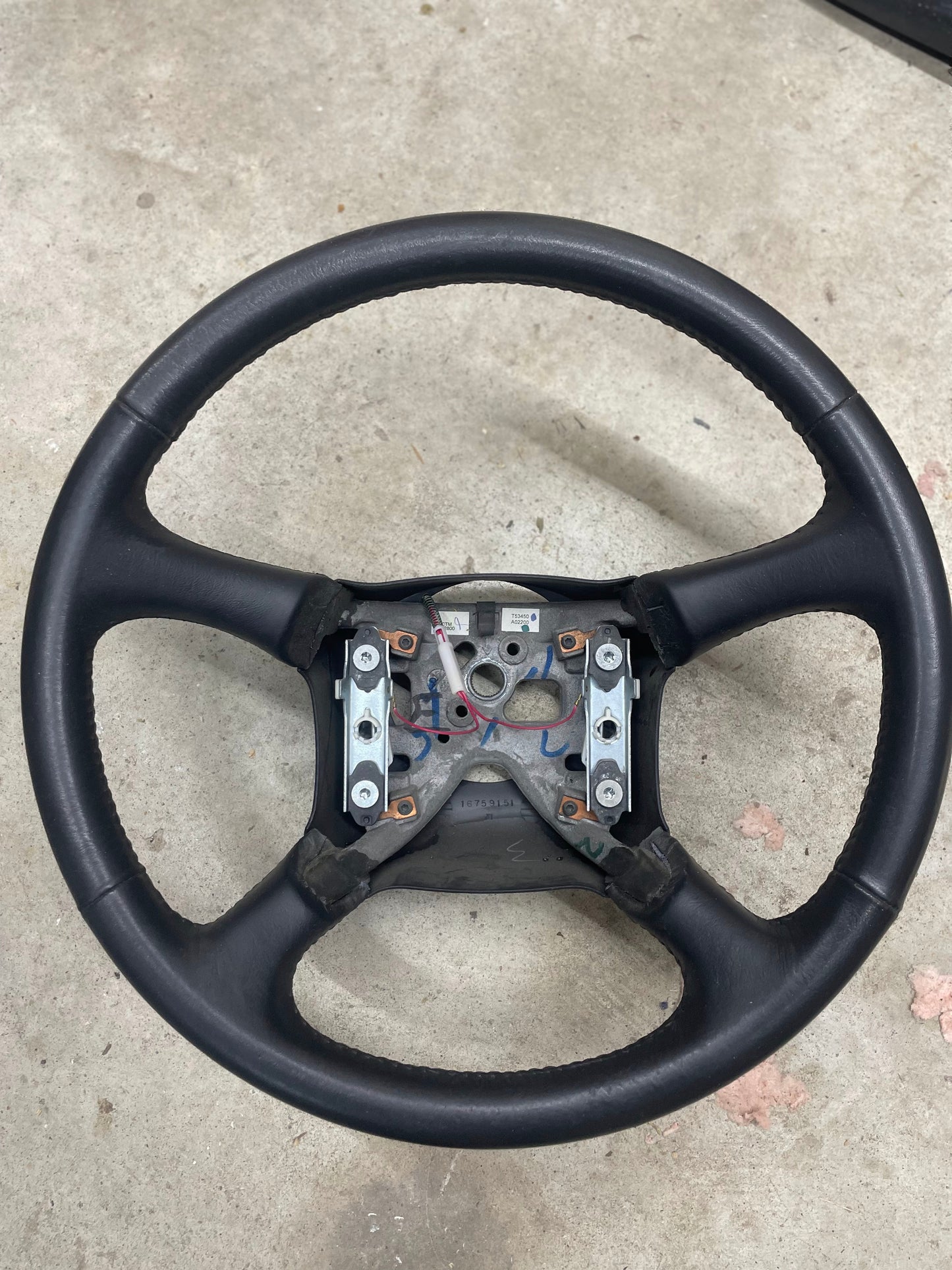 A General Motors OEM Leather Steering Wheel for 1999-2005 Chevy Silverado and S10 rests on a concrete floor, missing its center cover and revealing the metal mounting brackets and wiring inside.