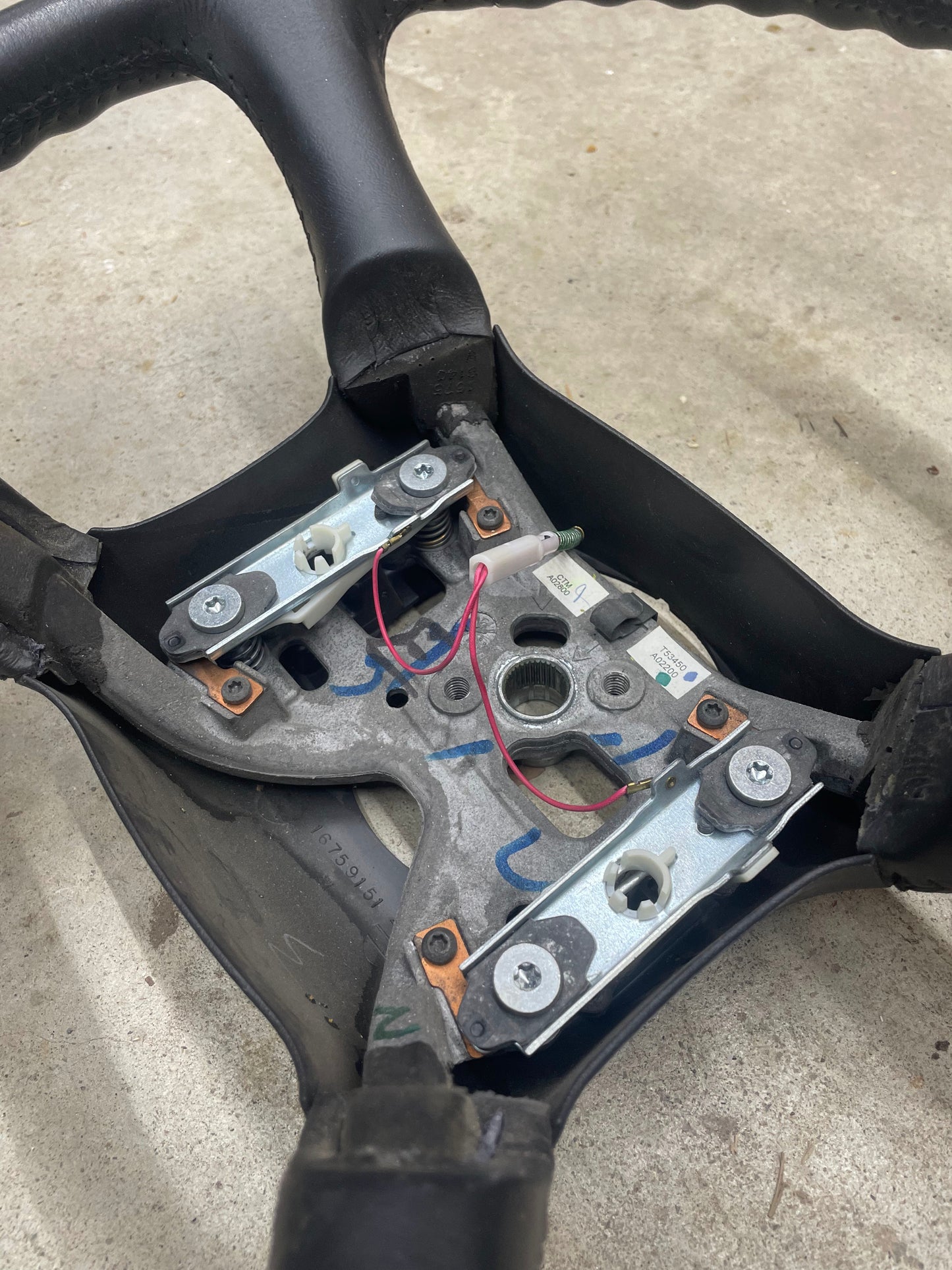 Close-up of the underside of a General Motors OEM Leather Steering Wheel for 1999-2005 Chevy Silverado, S10, etc., showing exposed metal parts and pink wires on concrete.