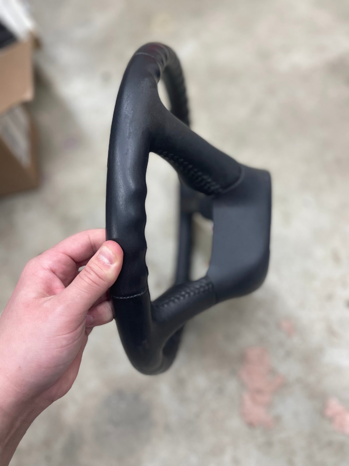 A hand grips an OEM Leather Steering Wheel for 1999-2005 GM Chevy Silverado S10 by General Motors, shown from the side with a contoured design against a blurred indoor background and concrete floor.