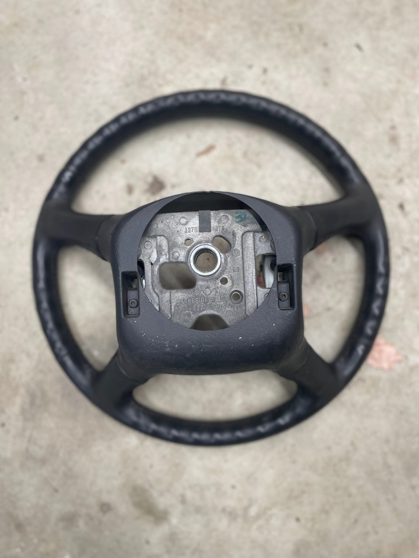 A General Motors OEM Leather Steering Wheel for 1999-2005 Chevy Silverado and S10 is shown without its center cover or airbag, exposing the inner metal and mounting parts on a concrete surface.