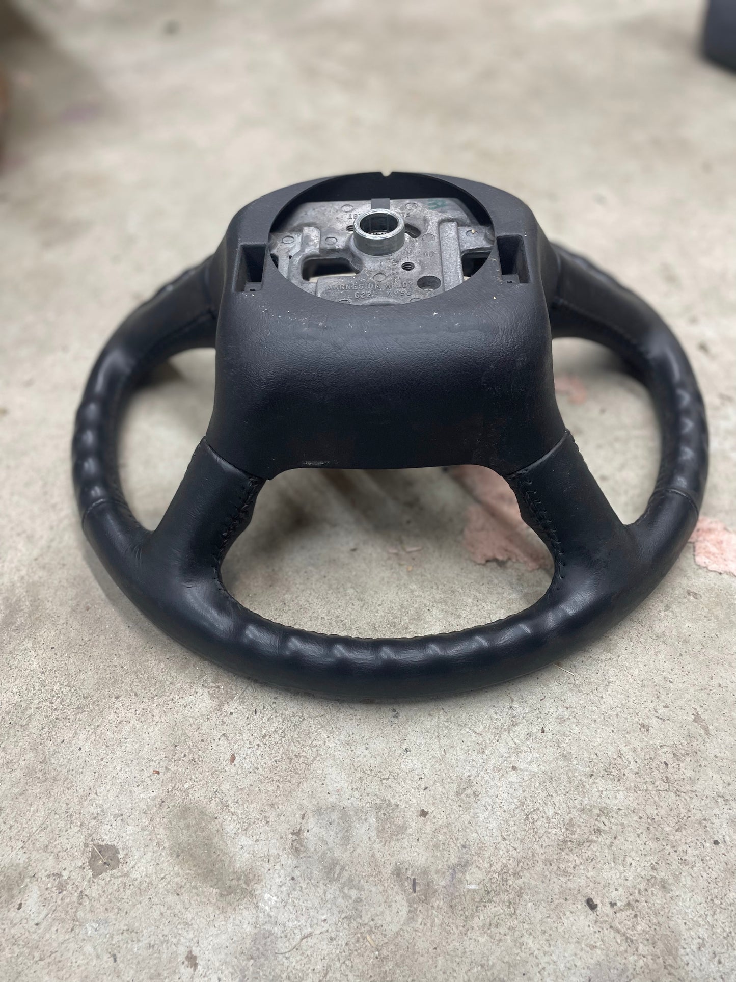 A General Motors OEM Leather Steering Wheel for 1999-2005 Chevy Silverado, S10, and more is shown without its center on a concrete floor, exposing the metal mounting area.