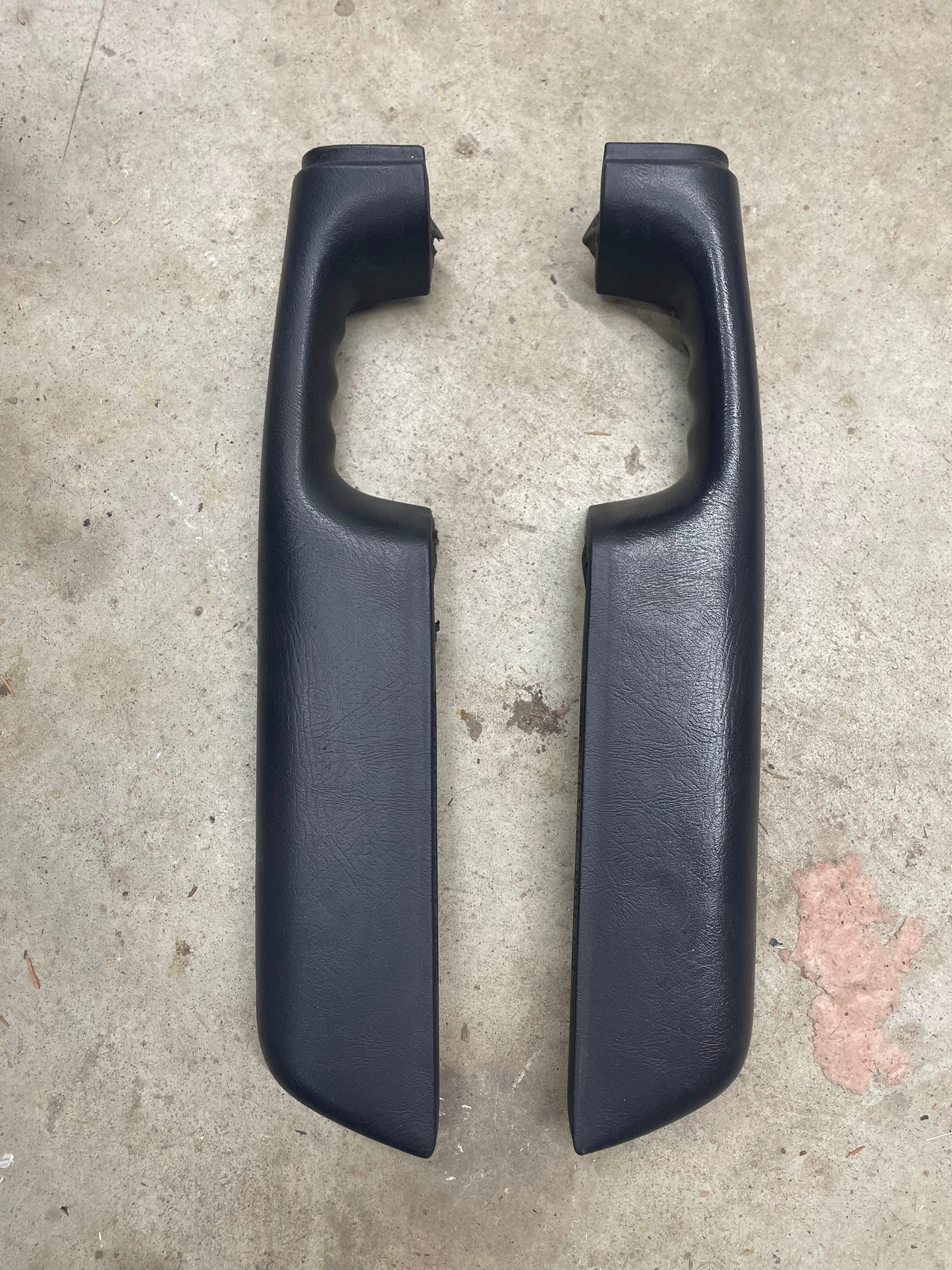 A pair of dark gray OEM General Motors front armrests, compatible with 1998–2005 Chevy S10 and Blazer, are placed side by side on concrete. The mirror-image handles feature finger grooves for grip.