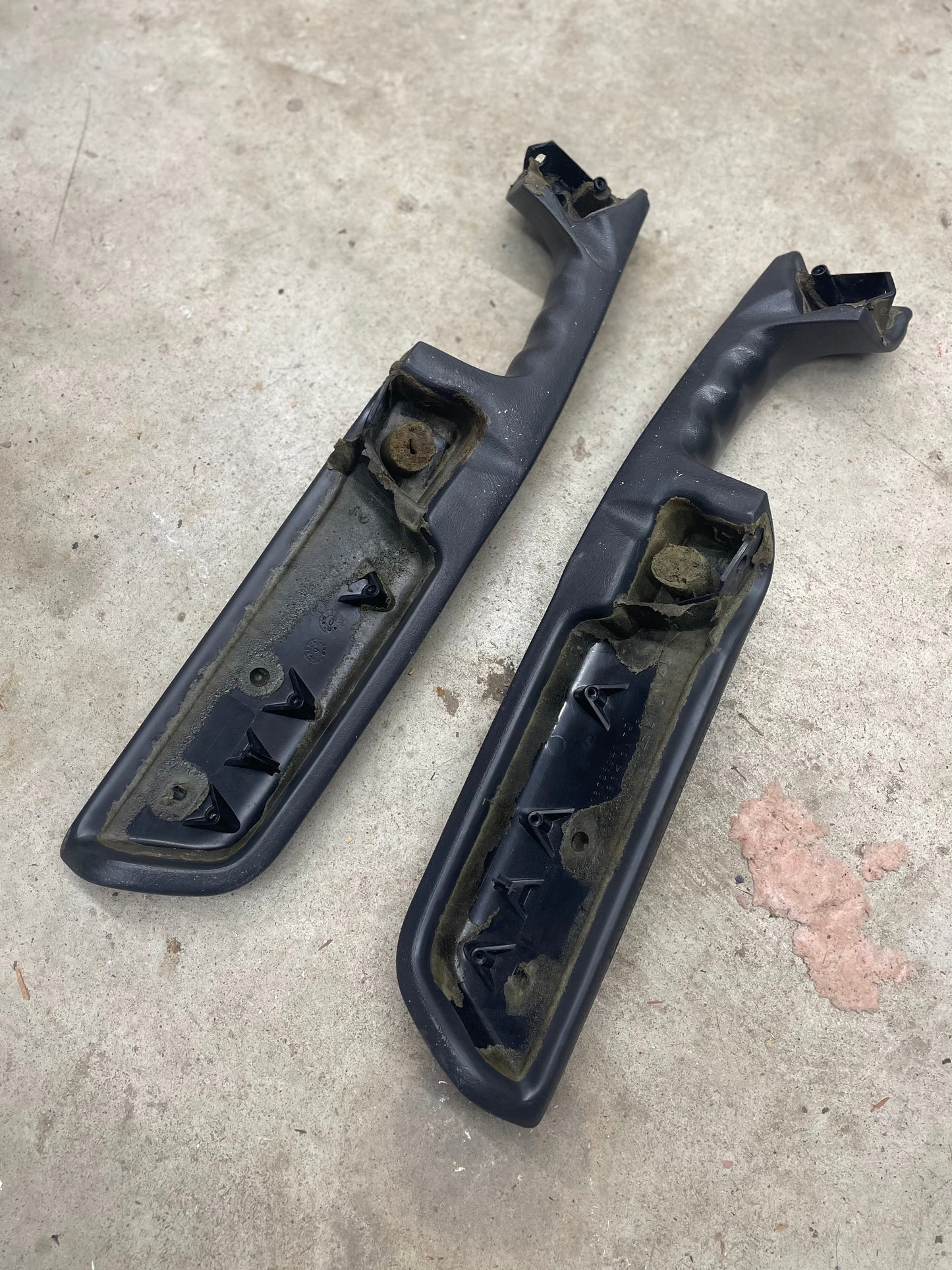 A pair of dark gray OEM front armrests by General Motors for 1998–2005 Chevy S10, Blazer, and more are lying on a concrete surface, showing wear, grime, rust, and broken mounting points.