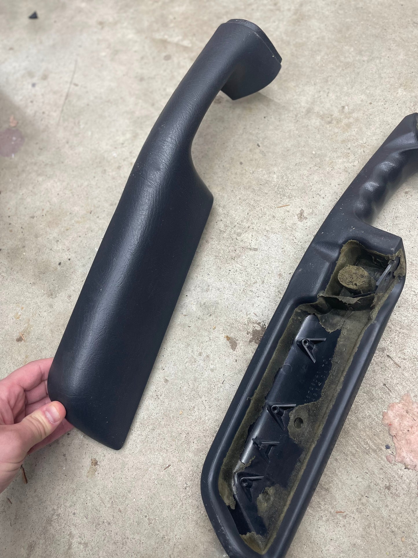 A hand holds a General Motors OEM front armrest cover in dark gray for 1998–2005 Chevy S10, Blazer, etc., next to its matching pair flipped to show the textured underside and attachment points on a concrete surface.