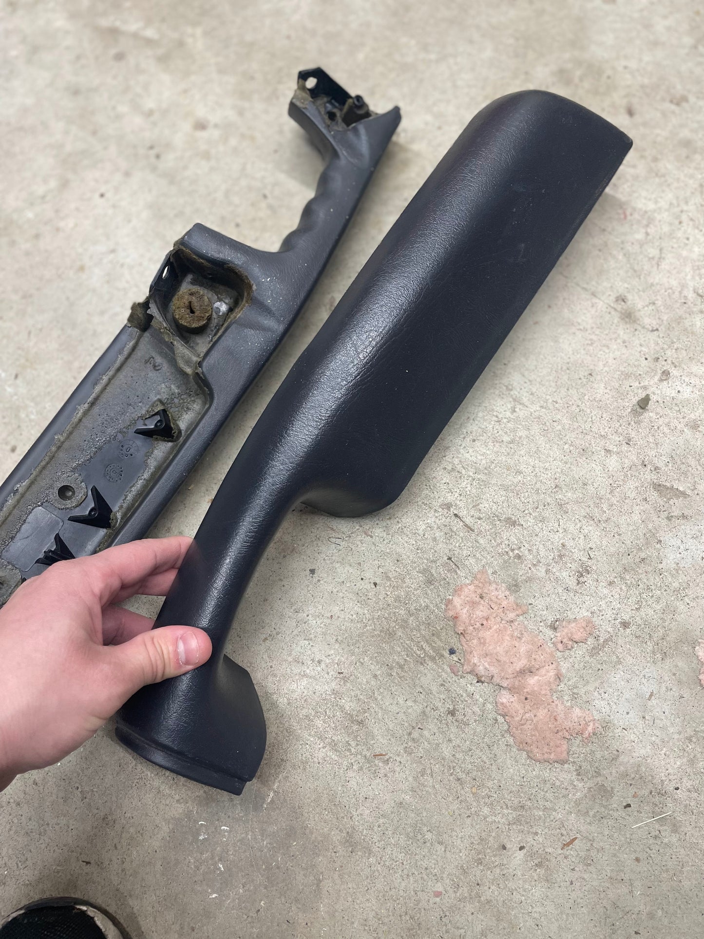 A hand holds a General Motors OEM front armrest in dark gray for 1998-2005 Chevy S10 and Blazer, next to a second, more worn piece lying on a concrete floor.