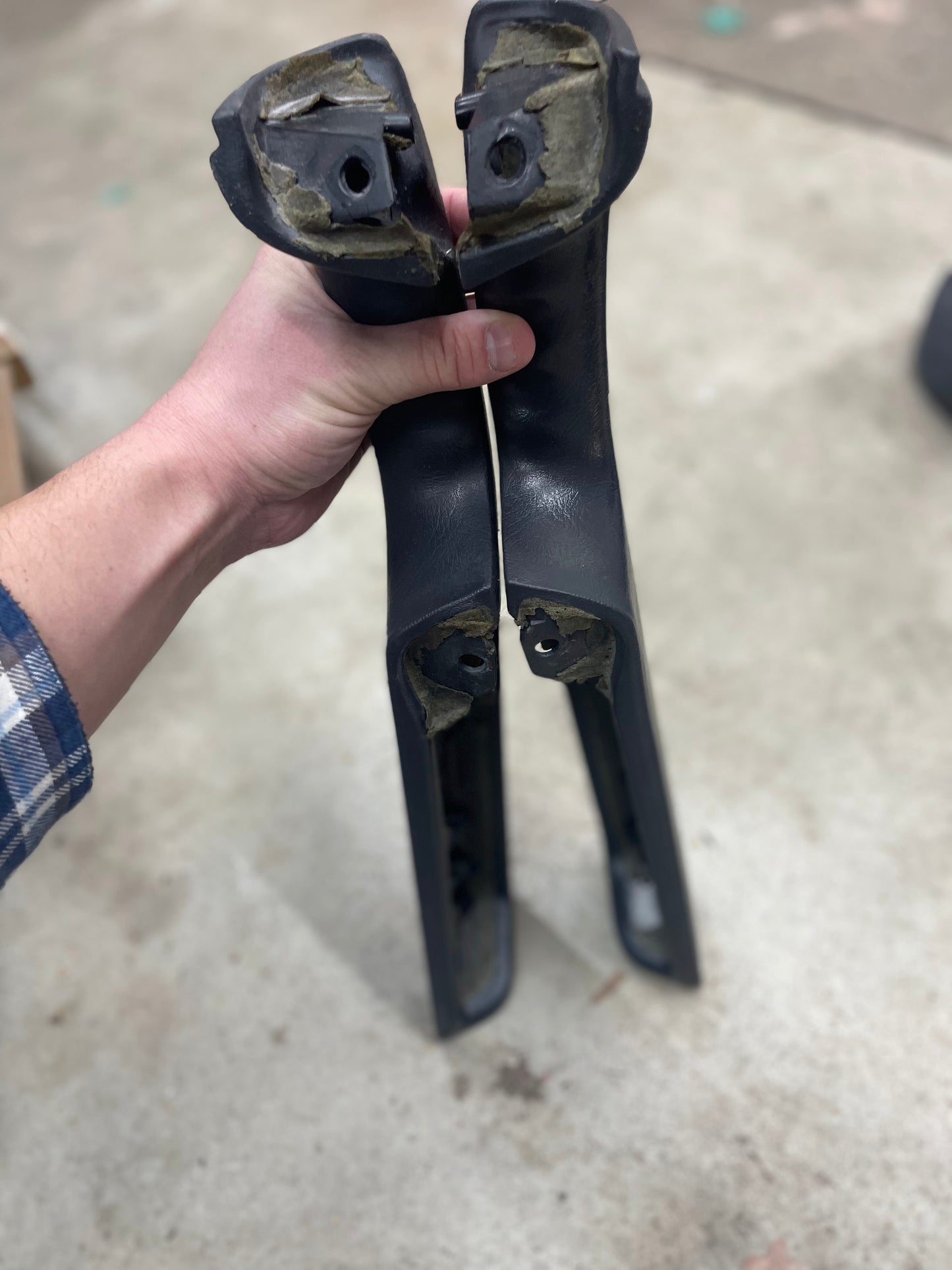 A person holds two broken General Motors OEM Front Armrest pieces in Dark Gray, made for 1998-2005 Chevy S10, Blazer, and more, displaying the damaged ends over a light-colored, slightly dirty floor.