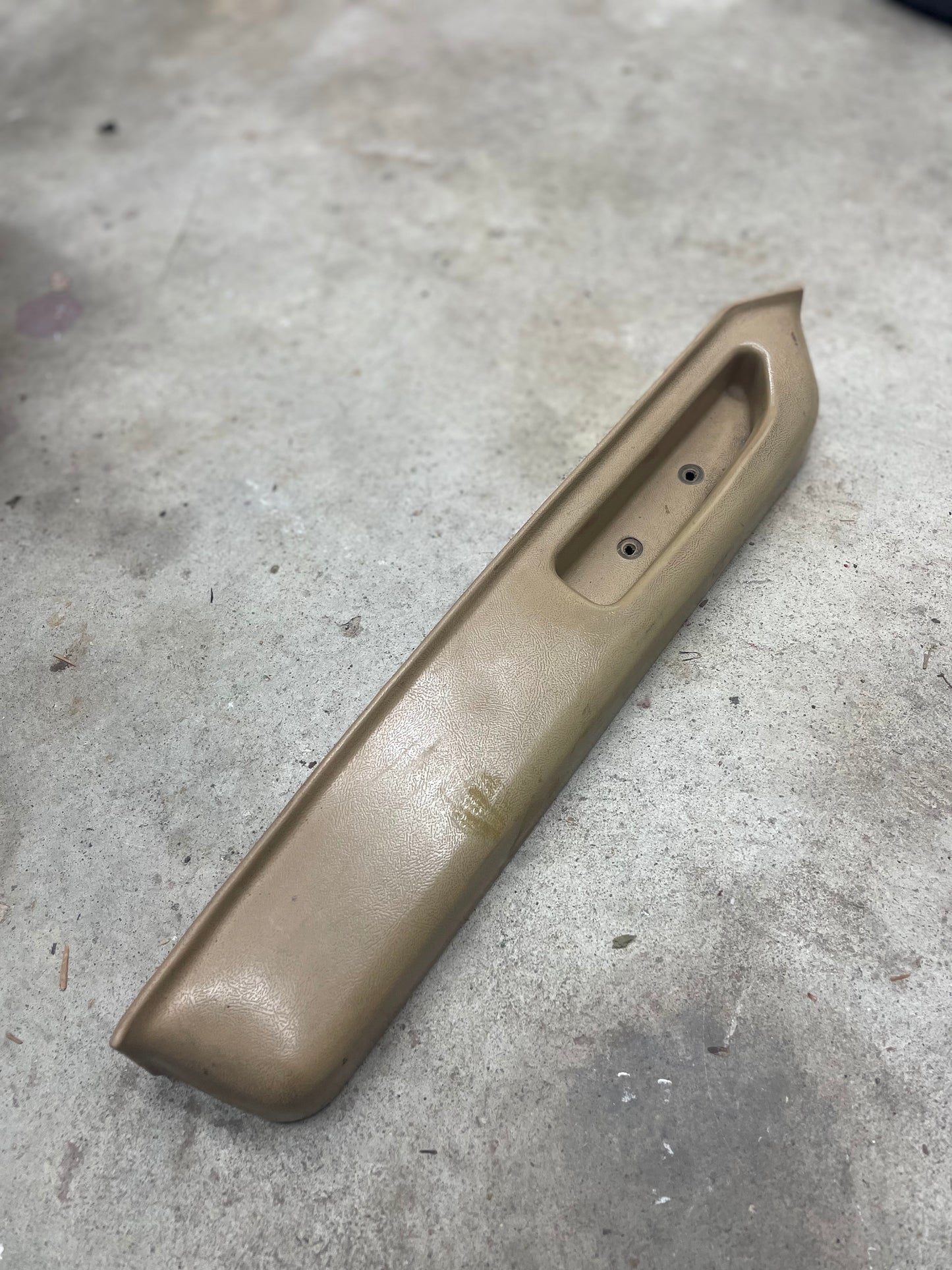 A General Motors OEM beige driver door panel armrest handle for 1986–1993 Chevy S10, Blazer, and more rests on a gray concrete floor, featuring a recessed area with two holes for mounting or attaching a handle or switch.