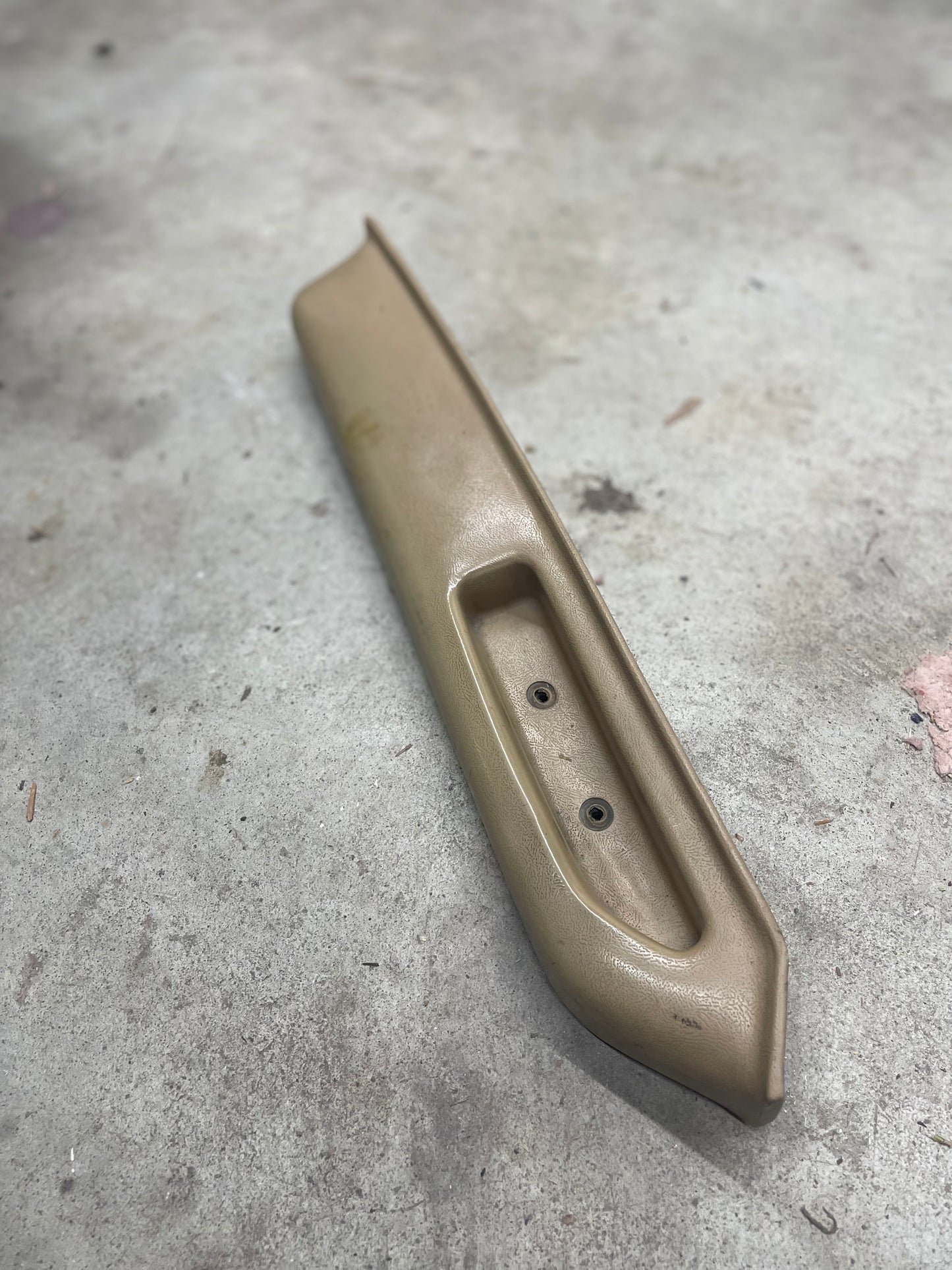 A General Motors OEM beige driver door panel armrest handle for 1986-1993 Chevy S10, Blazer, and more sits on a gray concrete floor; the armrest shows some wear and has a recessed area with two screw holes visible.