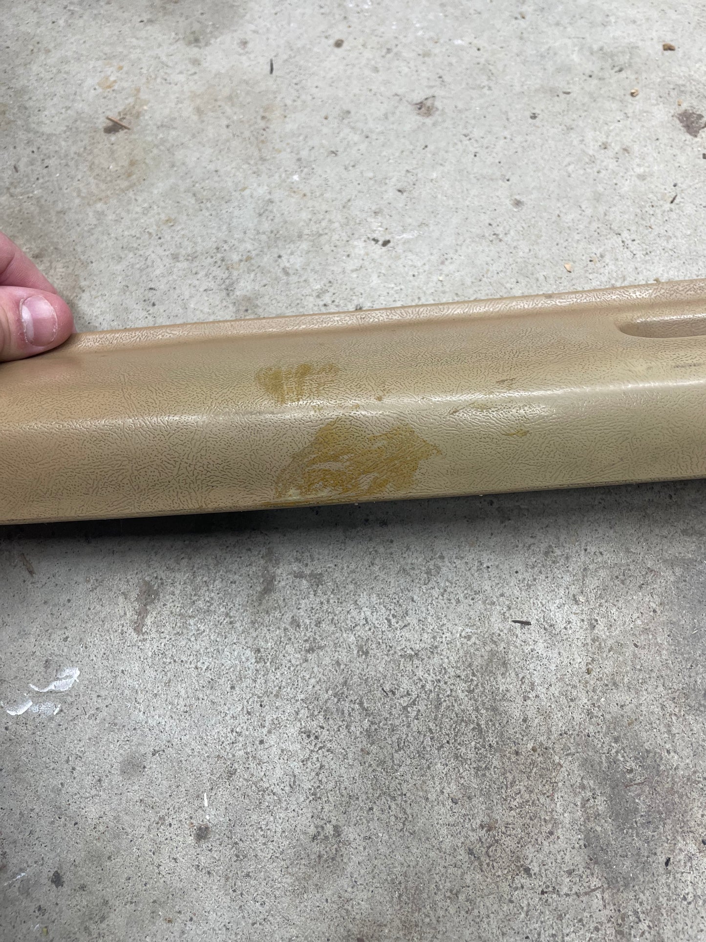 A hand holds the edge of a beige OEM driver door panel armrest handle by General Motors, made for 1986–1993 Chevy S10 and Blazer, showing a stain or discoloration on its plastic surface as it rests on a concrete floor.