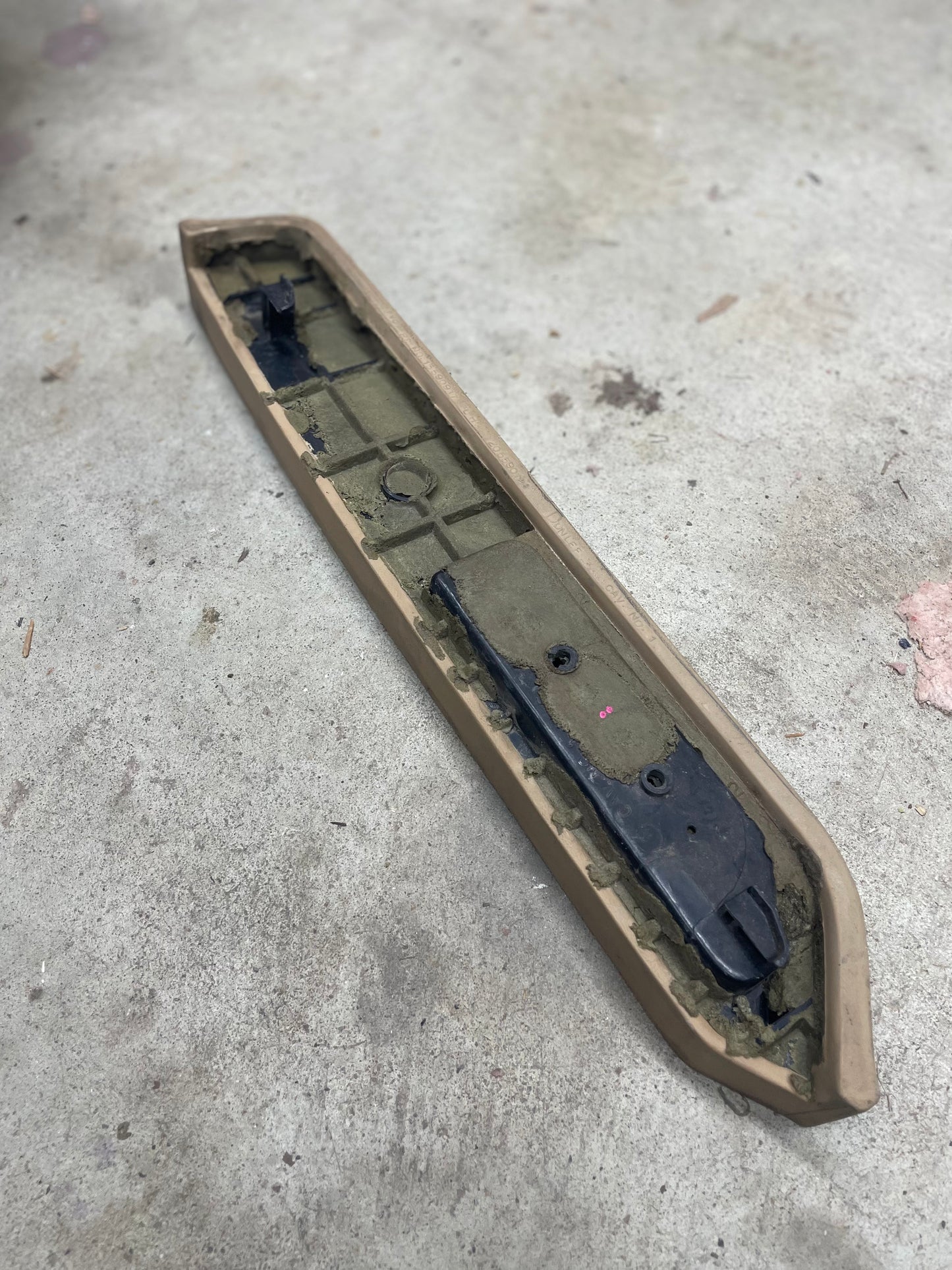 A close-up of a used OEM Driver Door Panel Armrest Handle in Beige for 1986-1993 Chevy S10/Blazer by General Motors rests on a concrete floor, showing slight dirt on its plastic and metal surface.