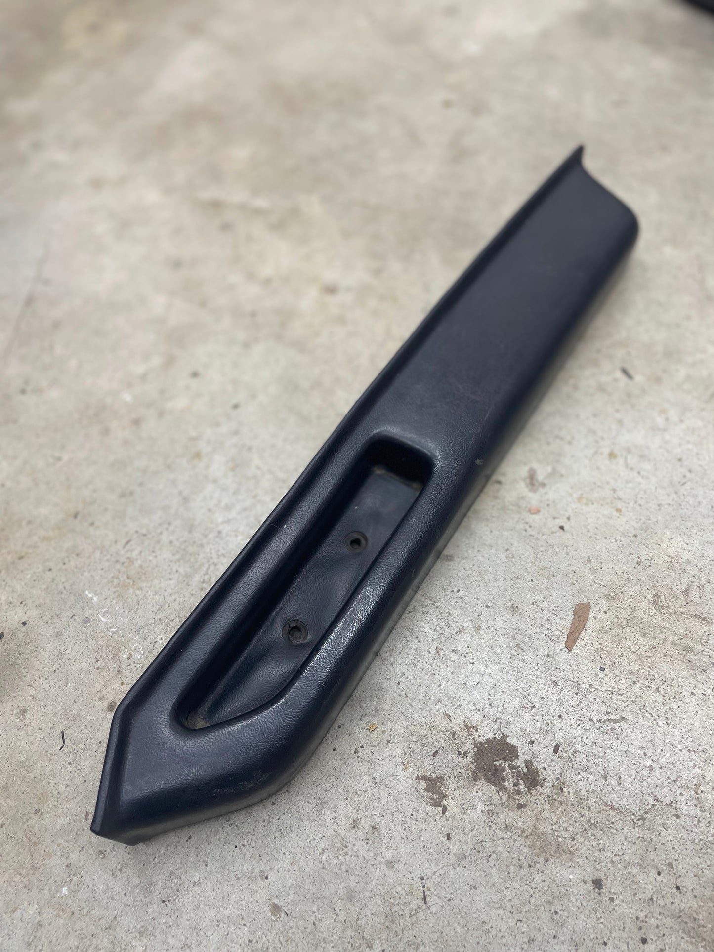 A black OEM General Motors passenger door panel armrest handle for 1986-1993 Chevy S10, Blazer, and GMC Jimmy lies on a light concrete floor; it features a recessed area with two screw holes and a pointed end—ideal as a replacement part.