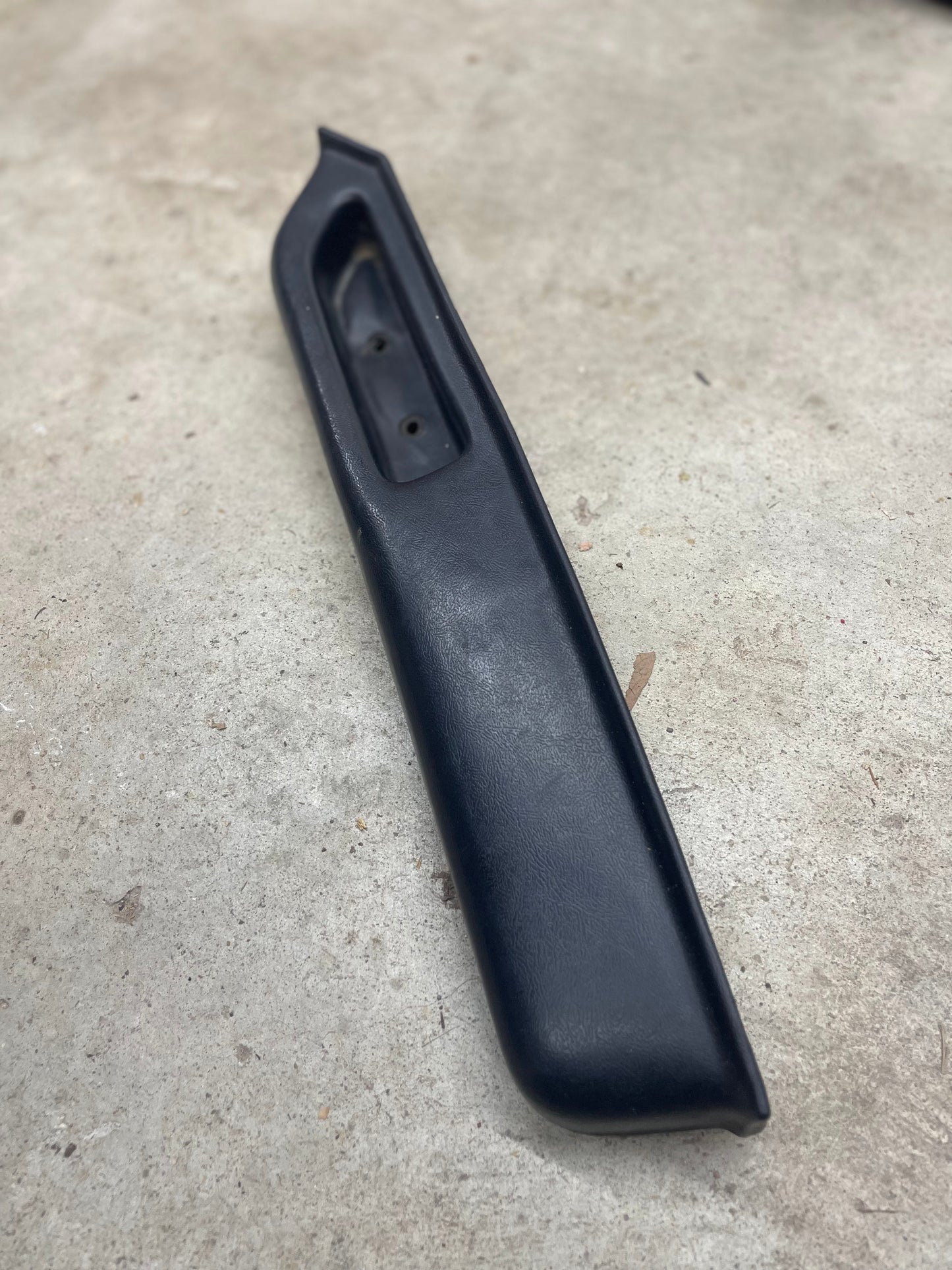 A black OEM Passenger Door Panel Armrest Handle by General Motors for 1986-1993 Chevy S10, Blazer, GMC Jimmy, and more is shown on a light textured concrete surface. The elongated trim piece features a recessed section and two mounting holes.
