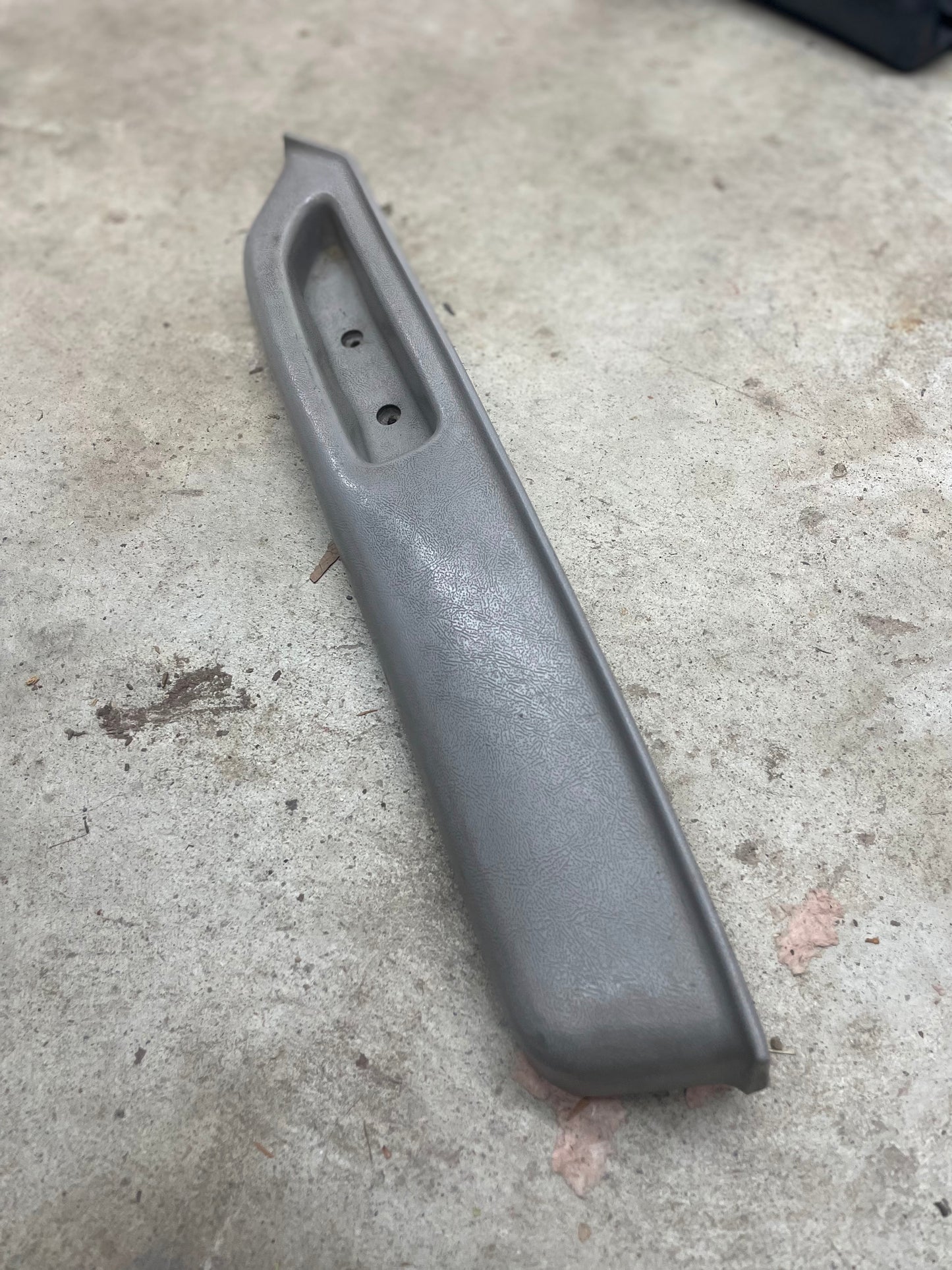 A General Motors OEM light gray passenger door panel armrest handle for 1986-1993 Chevy S10 Blazer, GMC Jimmy, and more lies on a concrete floor, showing two screw holes and a slightly curved design.