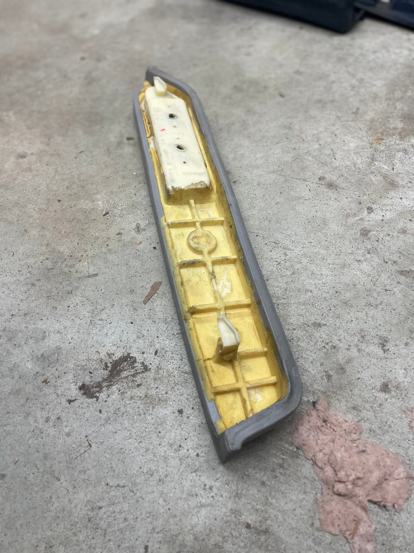A General Motors OEM Light Gray Passenger Door Panel Armrest Handle for 1986-1993 Chevy S10, Blazer, and GMC Jimmy rests on a concrete floor. The armrest shows ridges, mounting points, worn edges, and nearby debris.