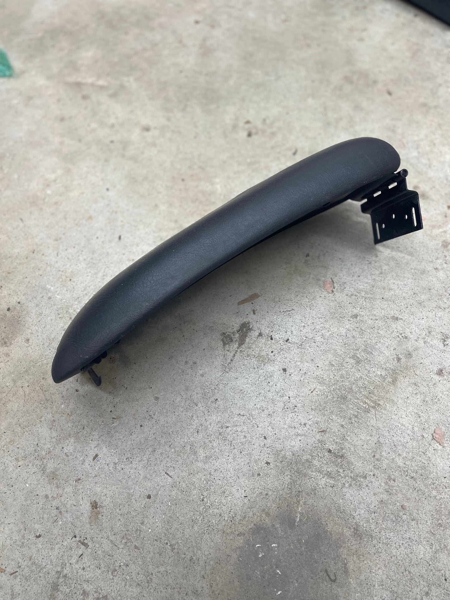 A dark gray OEM Center Console Lid Armrest by General Motors for 1998-2005 Chevy S10, Blazer, and more rests on rough light gray concrete, with a bracket attached to one end—ideal for replacing your original armrest.