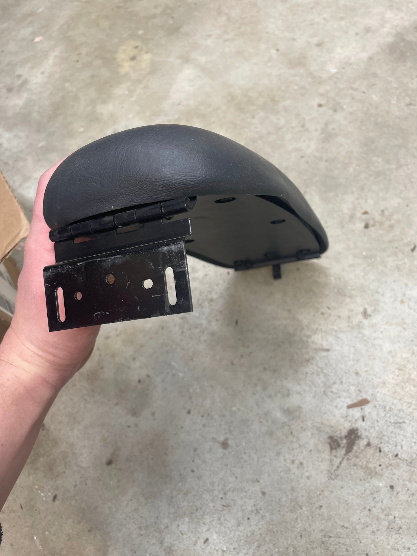 A hand holds a General Motors OEM Center Console Lid Armrest in dark gray for 1998-2005 Chevy S10 and Blazer, showing its hinge and bracket against a concrete floor.