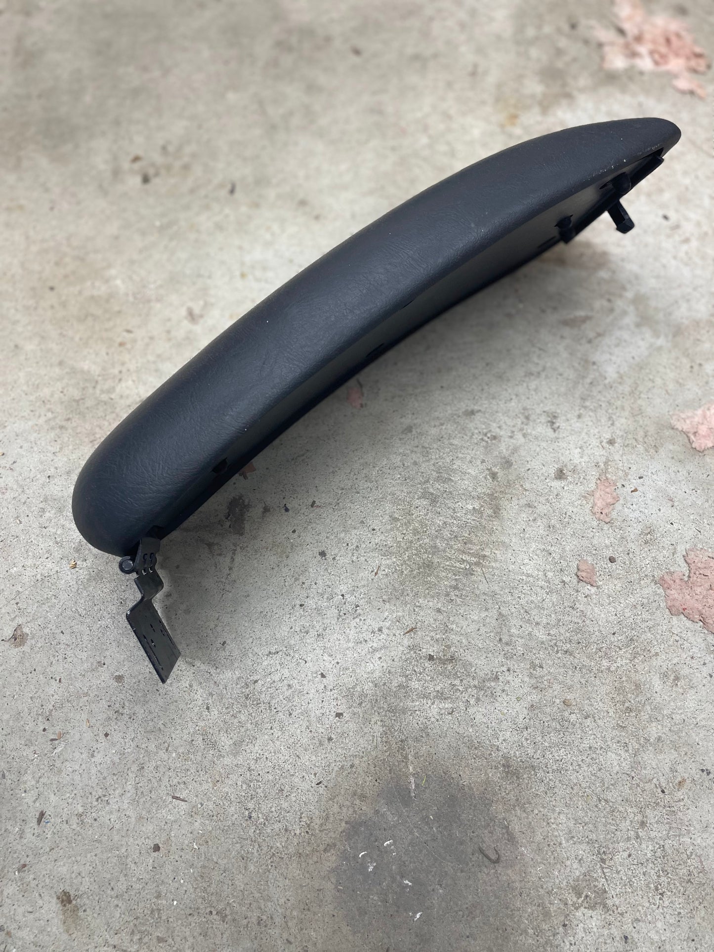 A dark gray OEM Center Console Lid Armrest by General Motors, with metal hinges, fits 1998-2005 Chevy S10/Blazer and more—ideal as a replacement for GM Graphite interiors. Shown on concrete with pink paint marks and light stains.