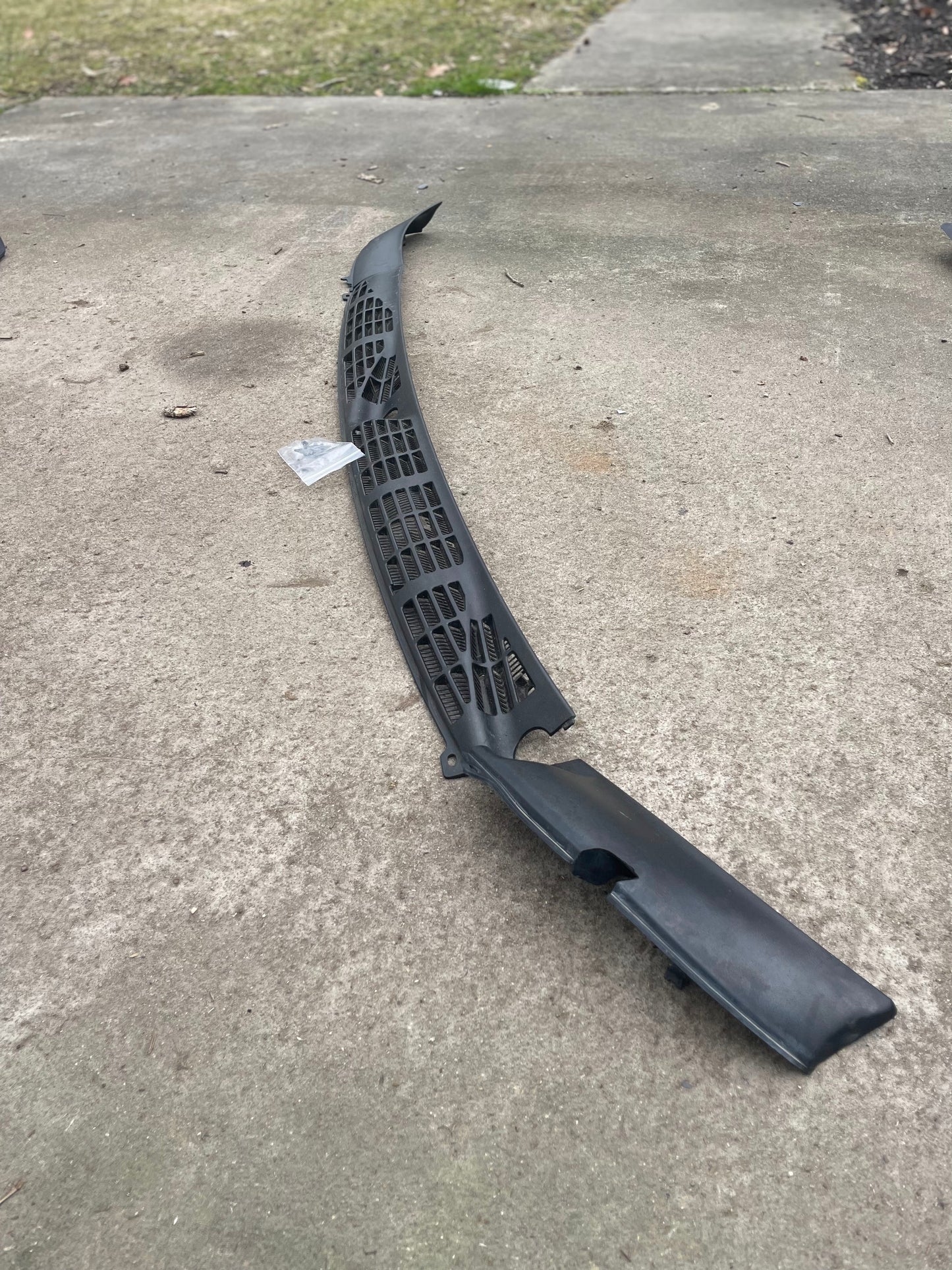 A General Motors OEM Wiper Cowl Cover Set with texture for 1994-2005 Chevrolet S10 and Blazer sits on a concrete driveway, with a lawn and sidewalk behind it. A small piece of paper is near the upper left side of the panel.