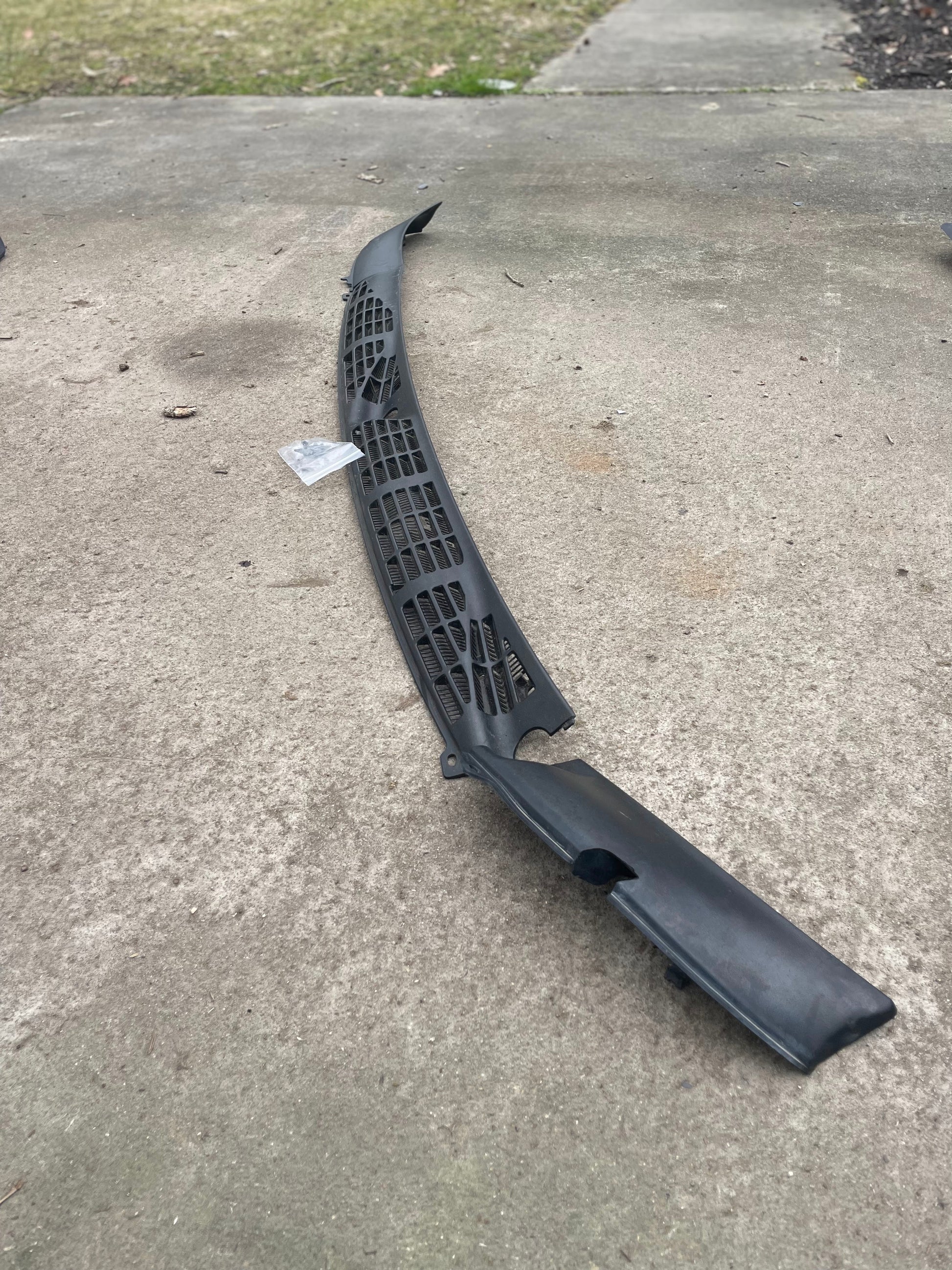 A General Motors OEM Wiper Cowl Cover Set with texture for 1994-2005 Chevrolet S10 and Blazer sits on a concrete driveway, with a lawn and sidewalk behind it. A small piece of paper is near the upper left side of the panel.